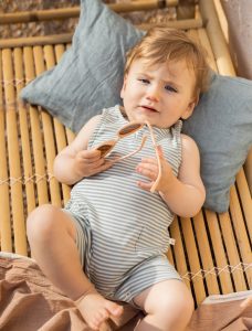 Bébé allongé portant une barboteuse sans manches rayée bleu vert 1+ in the Family, tenant une paire de lunettes de soleil.