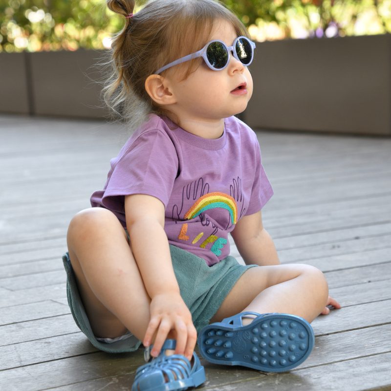 Petite fille assise portant un t-shirt violet Louise Misha, un short éponge bleu 1+ in the Family et des sandales Méduse bleu denim.