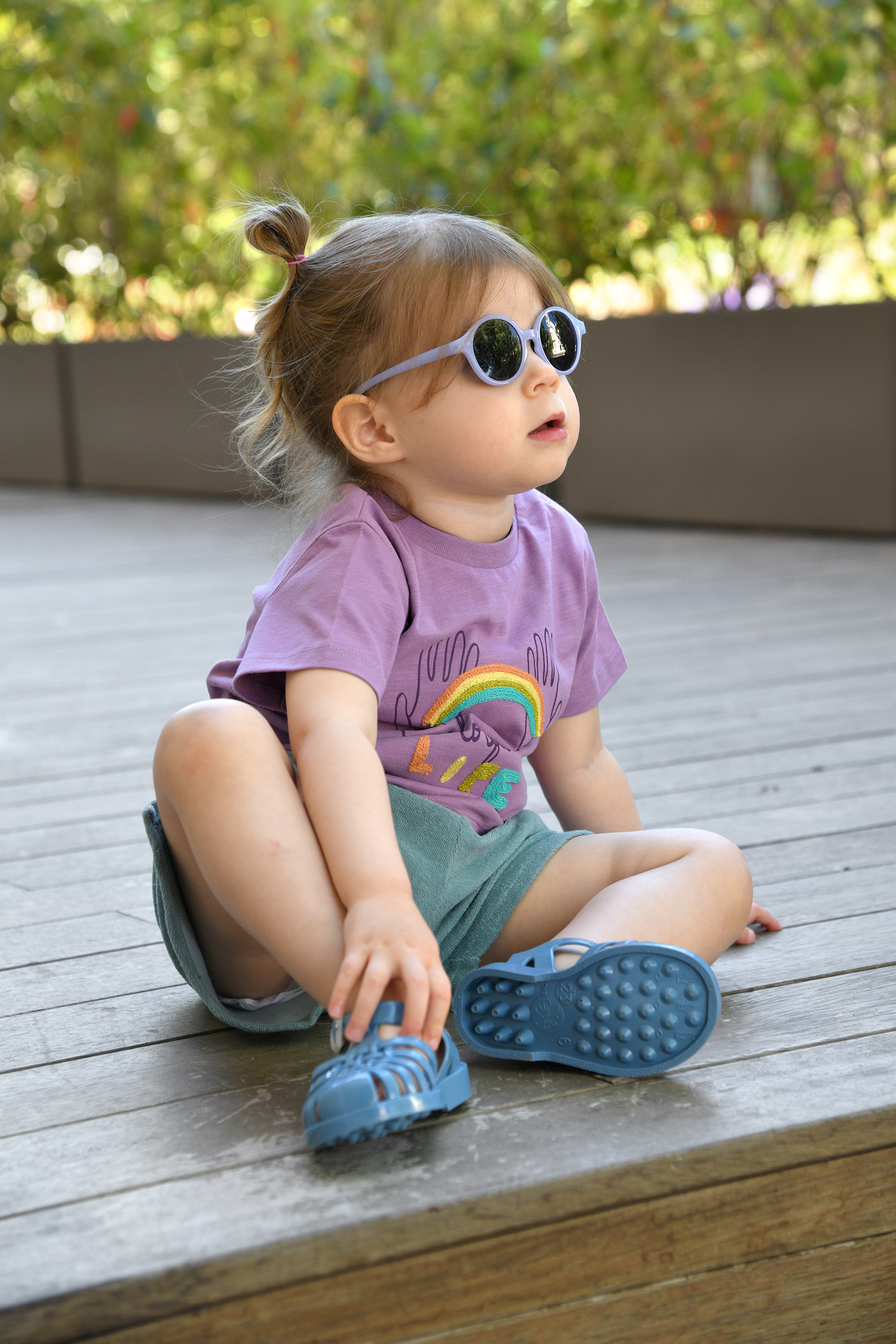 Petite fille assise portant un t-shirt violet Louise Misha, un short éponge bleu 1+ in the Family et des sandales Méduse bleu denim.