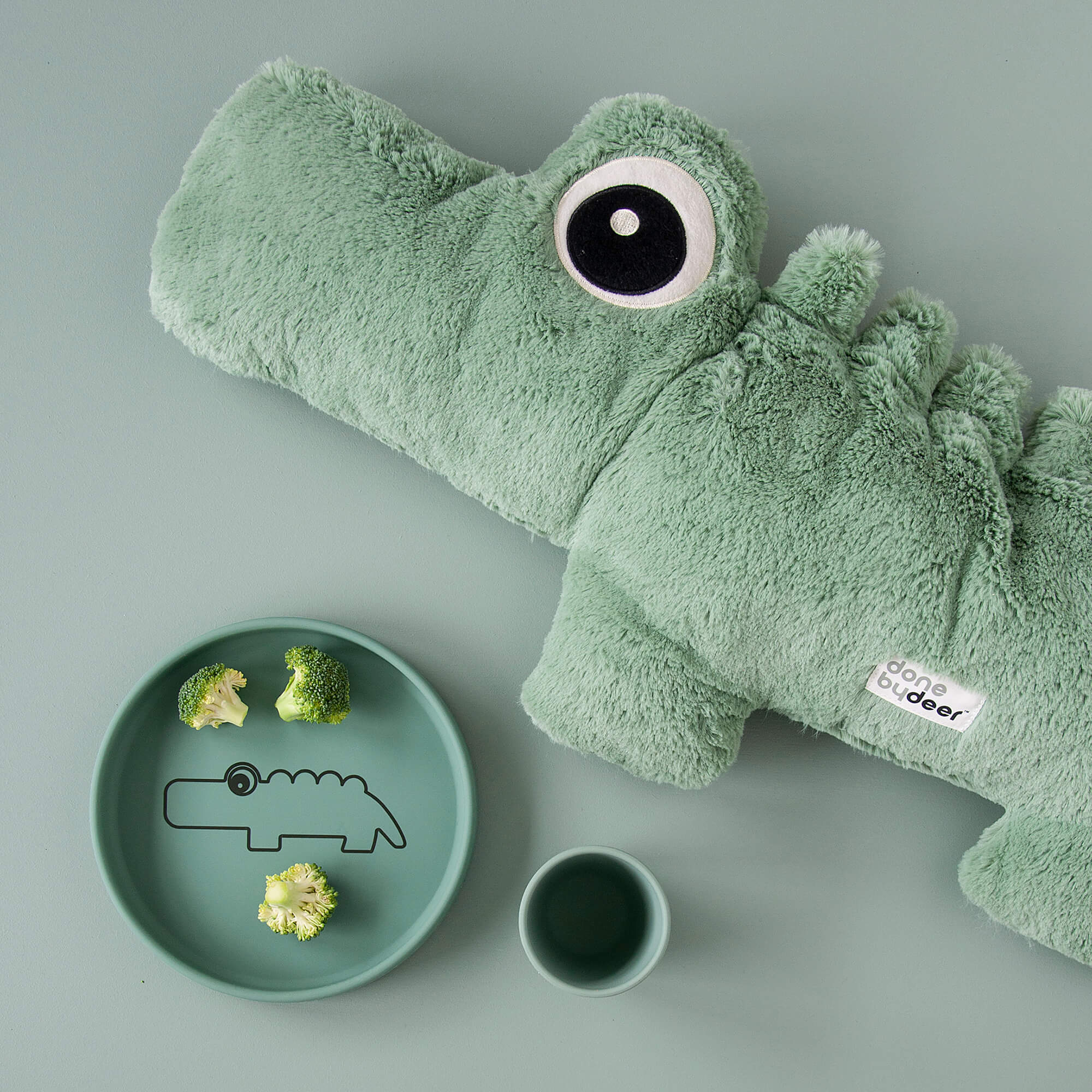 Peluche Croco verte Done by Deer à côté d’un bol en silicone assorti avec motif crocodile et des brocolis, verre à côté – ambiance douce et coordonnée