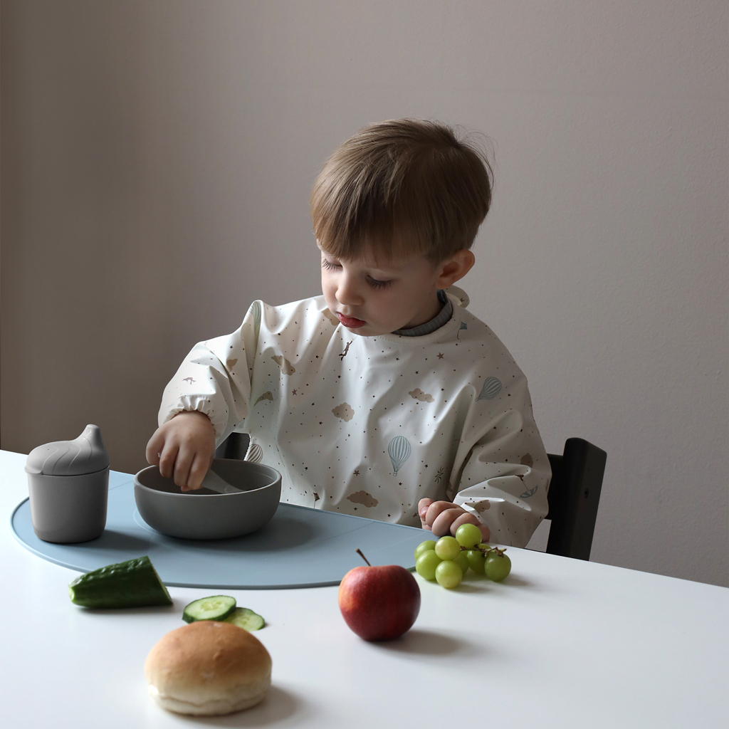 Enfant portant le bavoir à manches longues Dreamland Cam Cam pendant un repas, vaisselle grise et fruits sur la table.