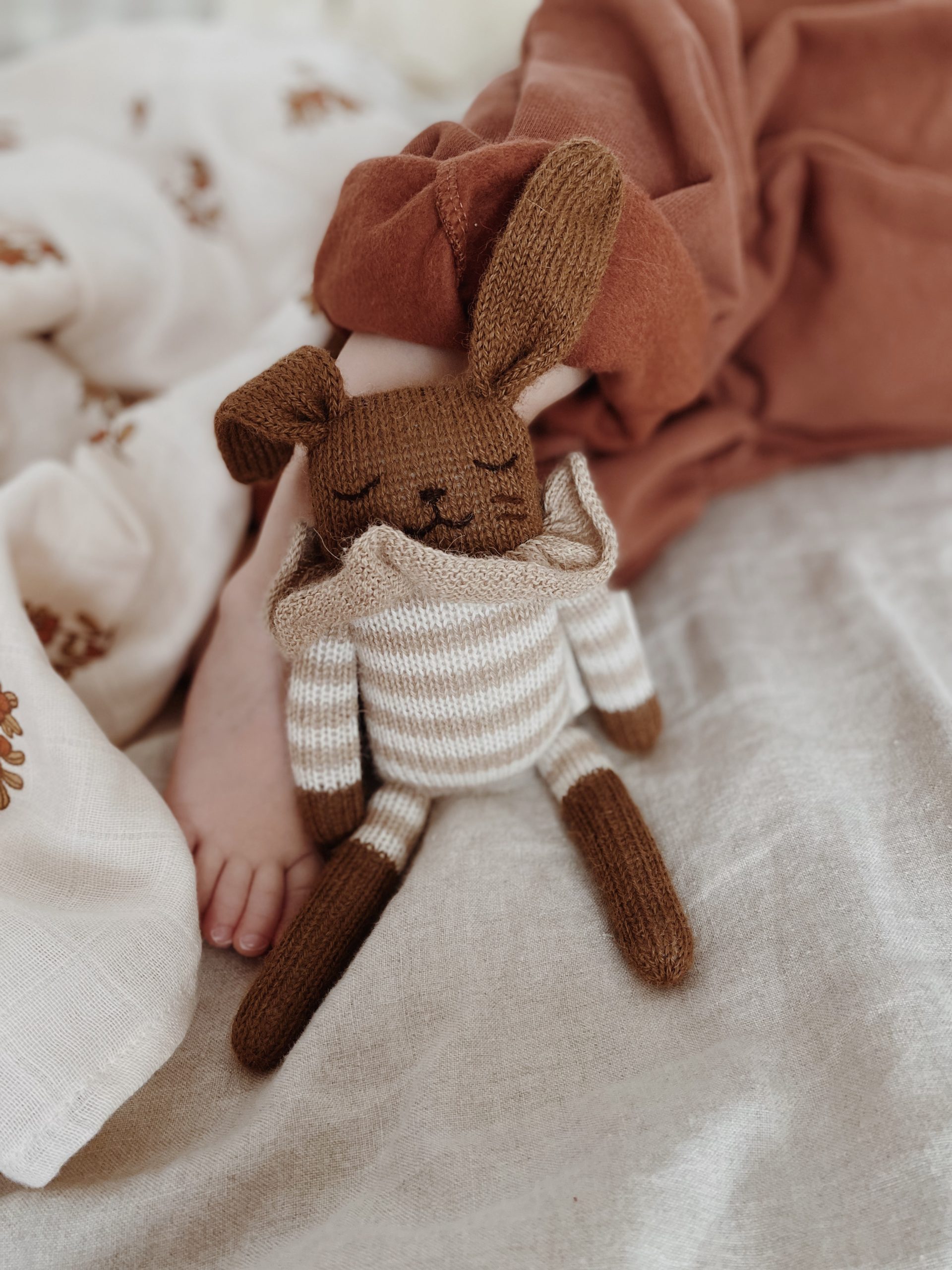 Doudou Lapin Combishort rayé sable Main Sauvage posé sur le pied d’un bébé allongé sur un lit – vue rapprochée