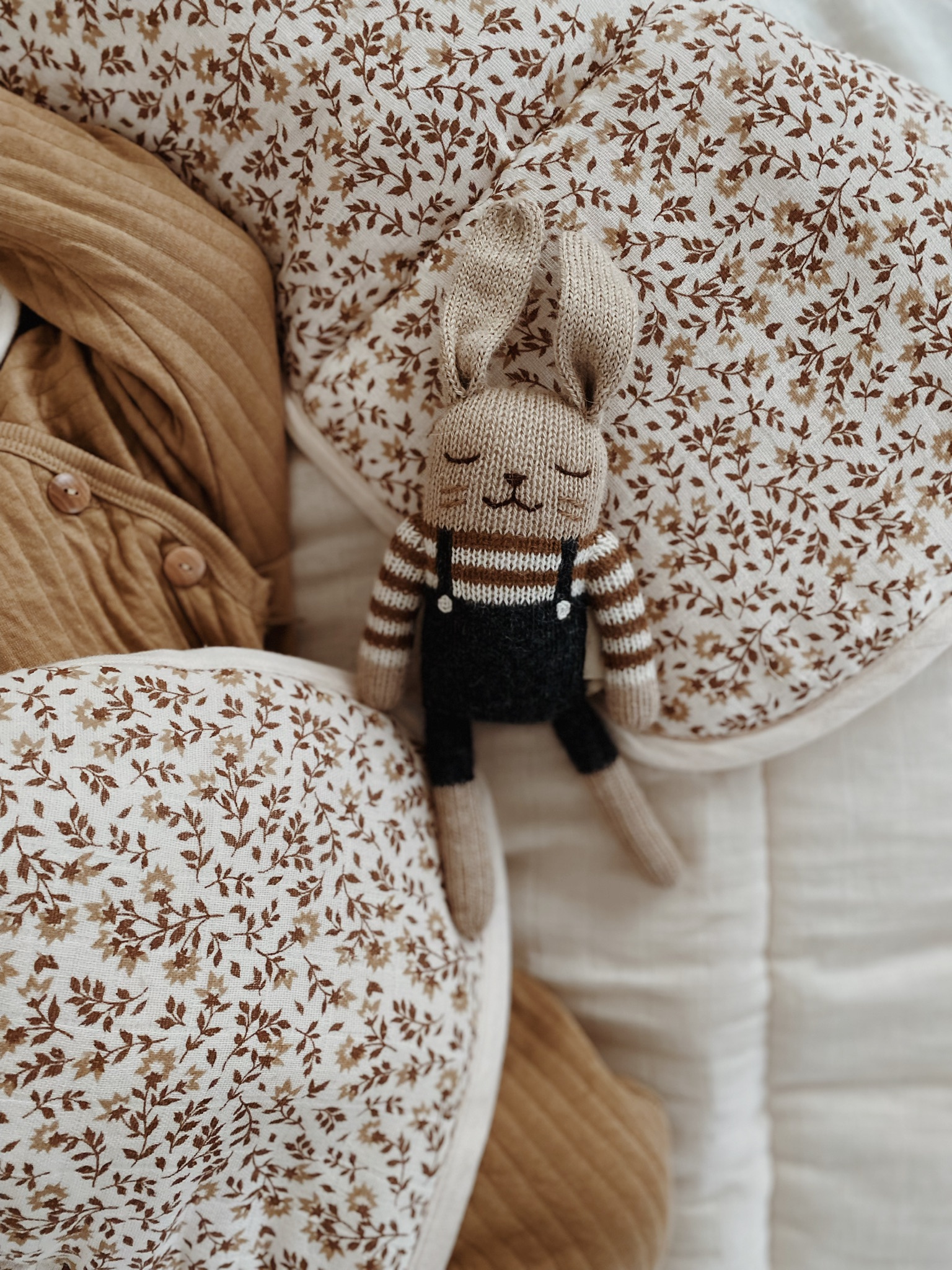 Doudou Lapin salopette noire Main Sauvage posé sur un lit avec une couette fleurie et une couverture – ambiance tranquille