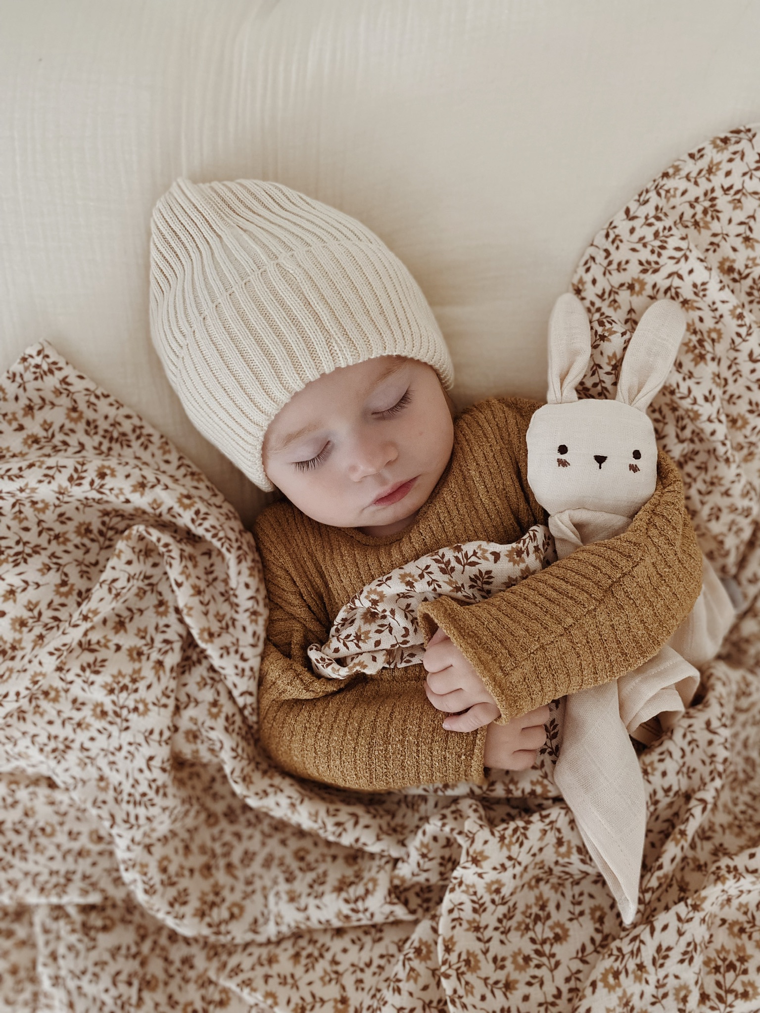 Bébé endormi vu du dessus avec le doudou lange Lapin écru Main Sauvage entre ses bras, drap fleuri, pull moutarde et bonnet crème
