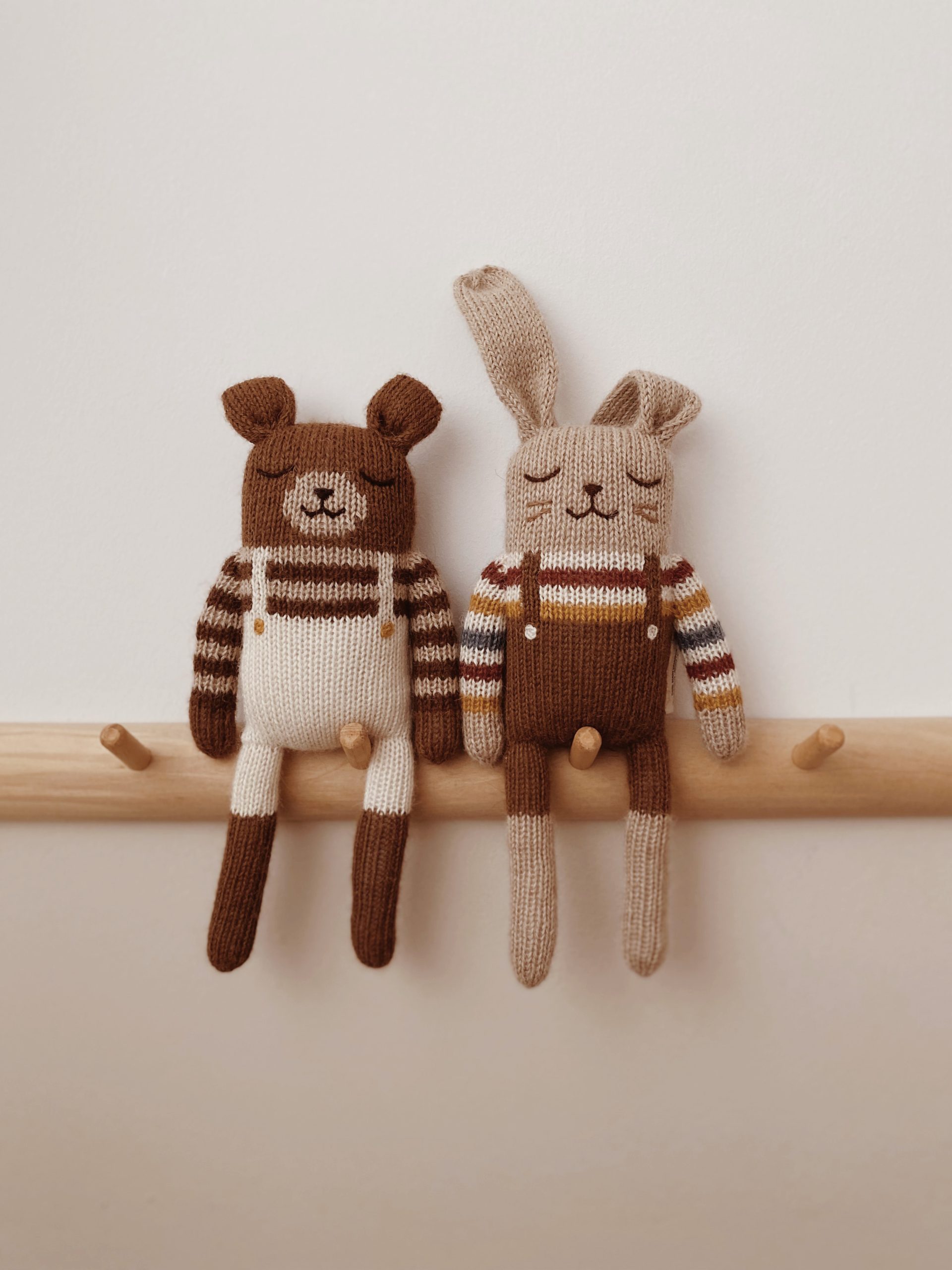 Doudou Lapin brun avec pull arc-en-ciel Main Sauvage et doudou Ourson assorti accrochés sur un porte-vêtements mural – ambiance complice