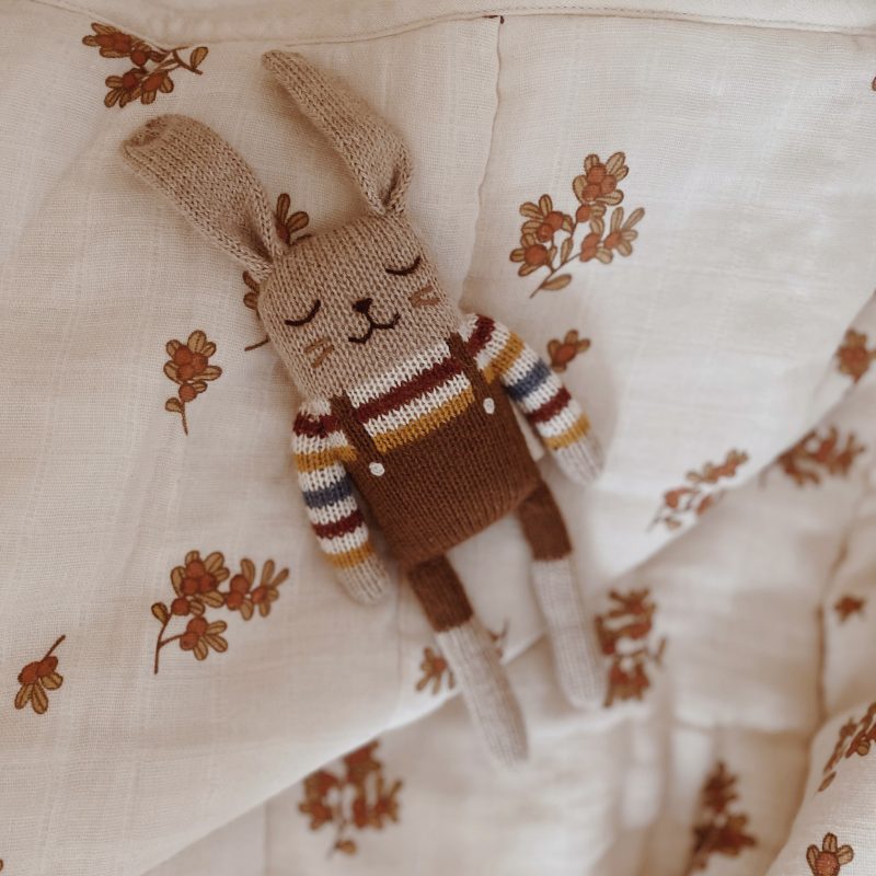 Doudou Lapin brun avec pull arc-en-ciel Main Sauvage posé sur un lange à motifs fleuris