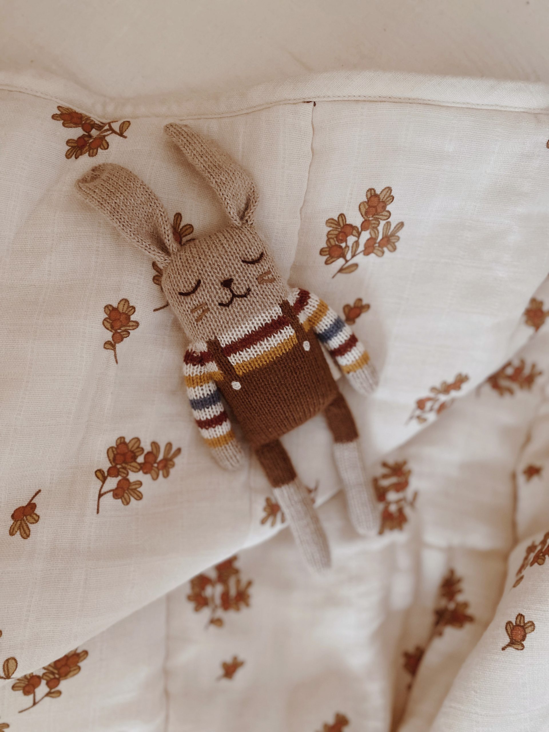Doudou Lapin brun avec pull arc-en-ciel Main Sauvage posé sur un lange à motifs fleuris
