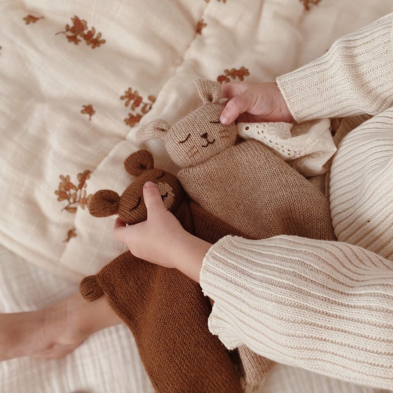 Doudous plats Lapin sable et noisette Main Sauvage tenus côte à côte dans les mains d’un bébé allongé sur un lit – vue rapprochée