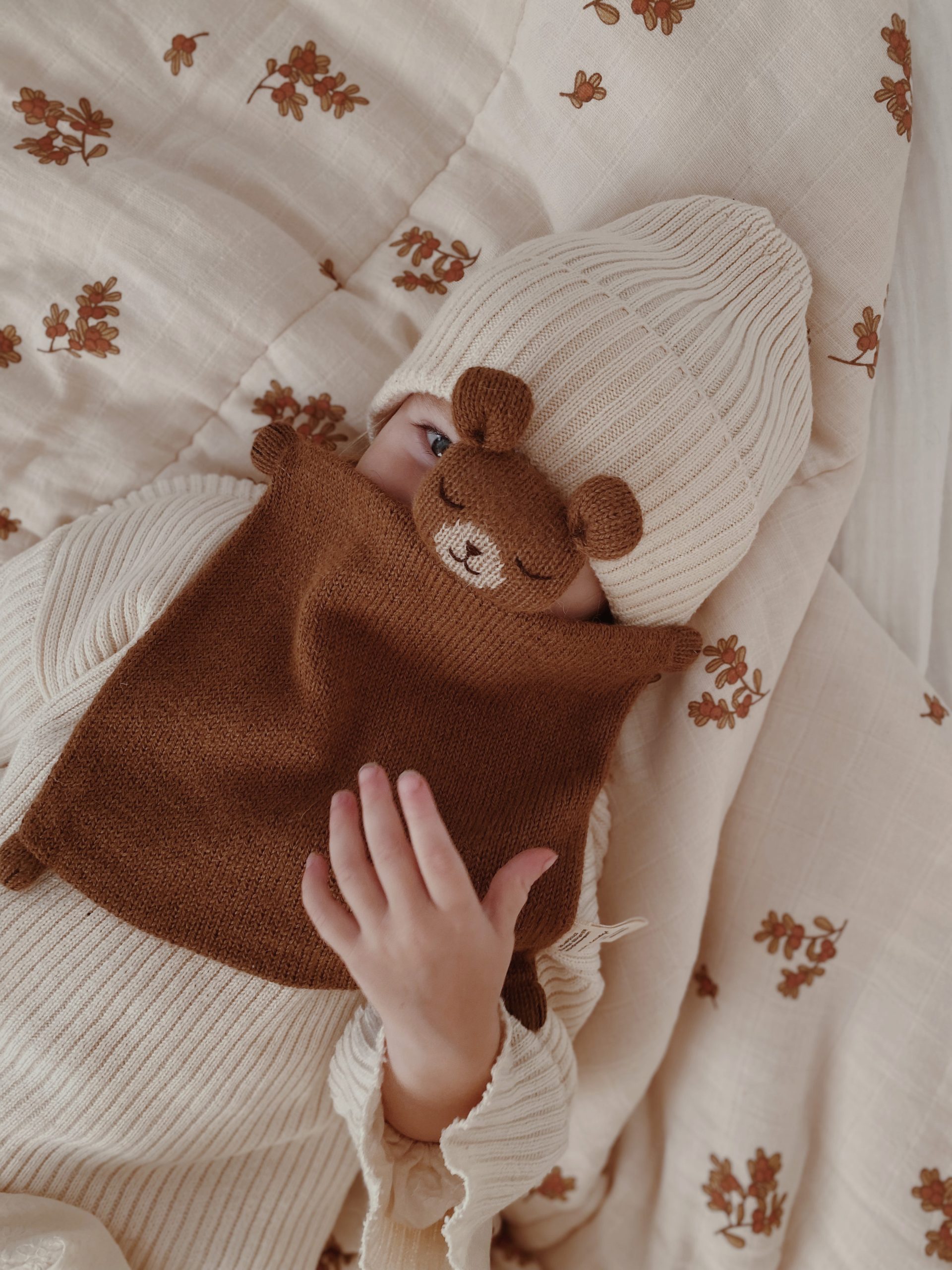 Bébé allongé vu de face tenant le doudou plat Ourson noisette Main Sauvage devant son visage, les yeux tournés vers l’objectif