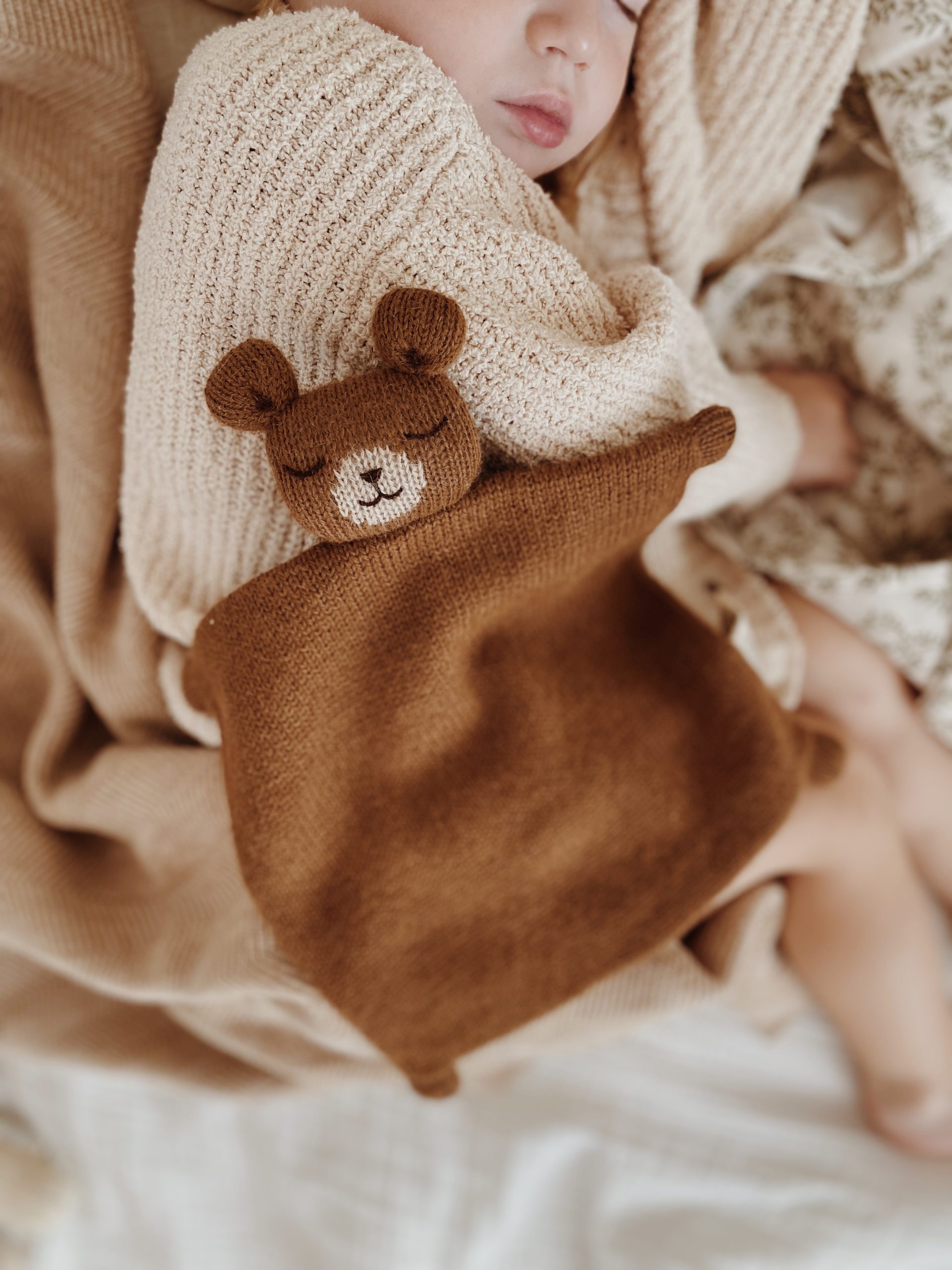 Bébé endormi de profil sur un lit avec le doudou plat Ourson noisette Main Sauvage posé sur lui – ambiance paisible