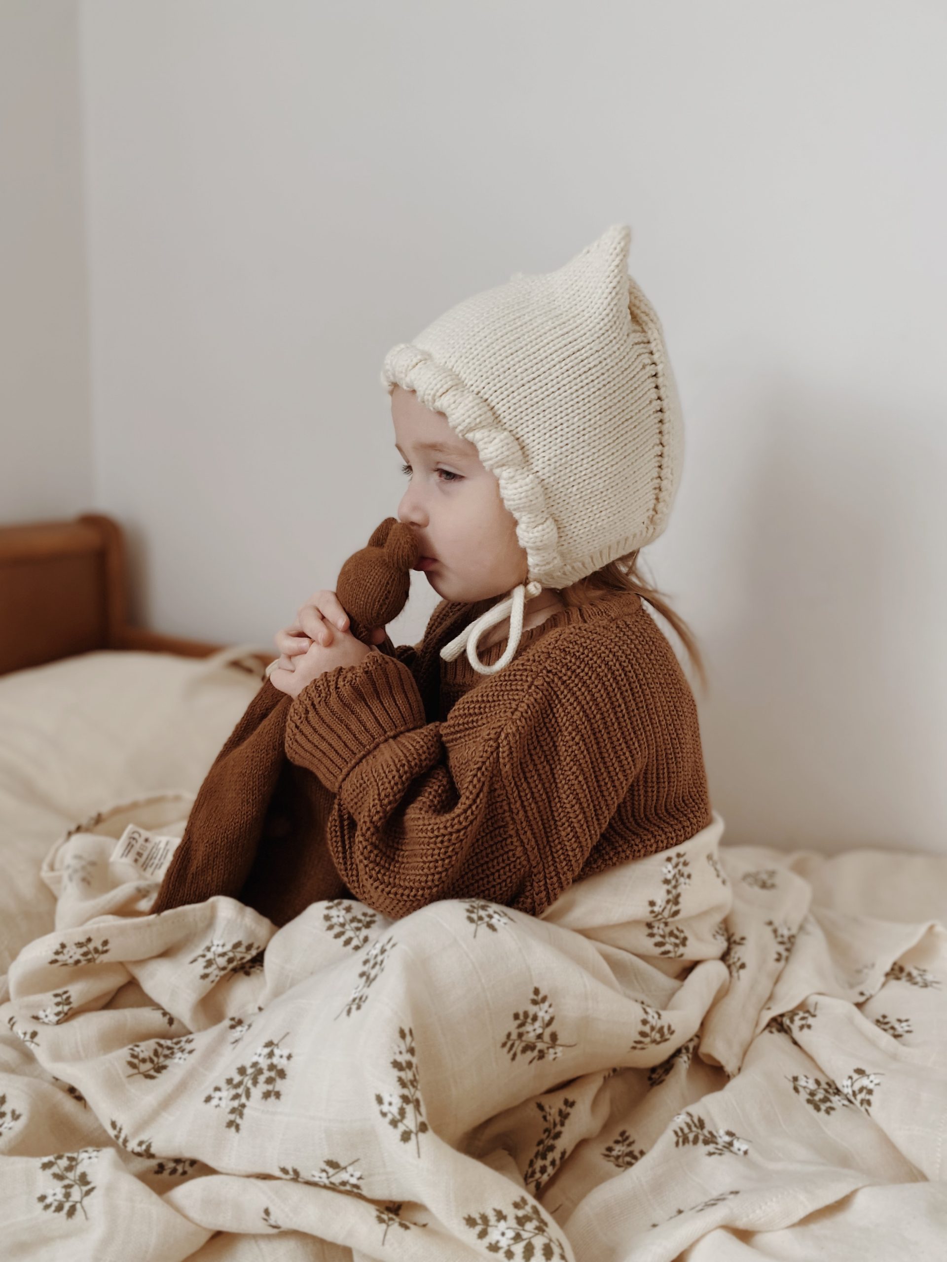 Enfant assis sur un lit avec un bonnet beige à oreilles, tenant le doudou plat Ourson noisette Main Sauvage contre sa bouche pour lui faire un bisou – vue de profil
