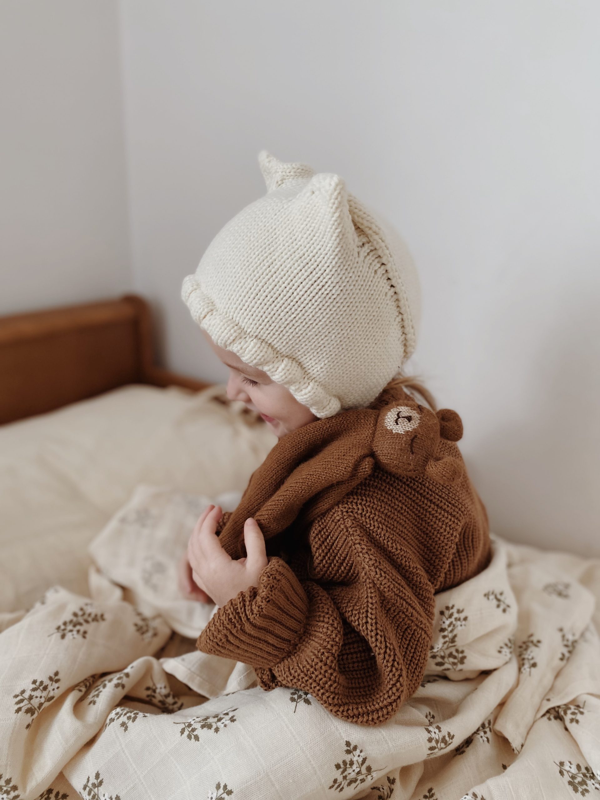 Enfant assis sur un lit, vu de profil et de dos, portant un bonnet beige à oreilles et souriant avec le doudou plat Ourson noisette Main Sauvage posé sur son épaule – ambiance joyeuse et douce