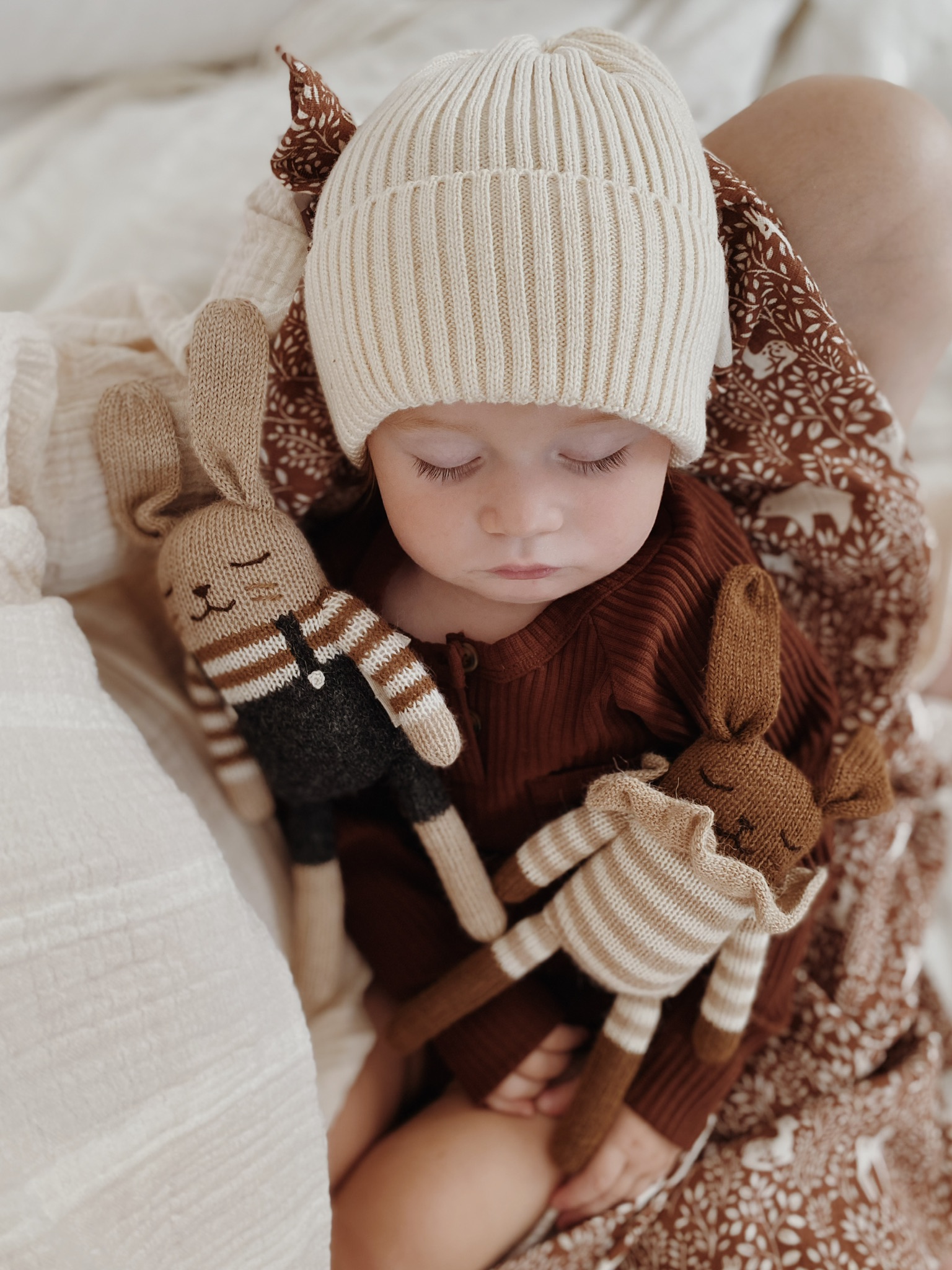 Bébé endormi sur les jambes de sa mère avec le doudou Lapin Combishort rayé sable Main Sauvage et un autre doudou lapin en salopette posés sur lui – sur un lit avec langes autour