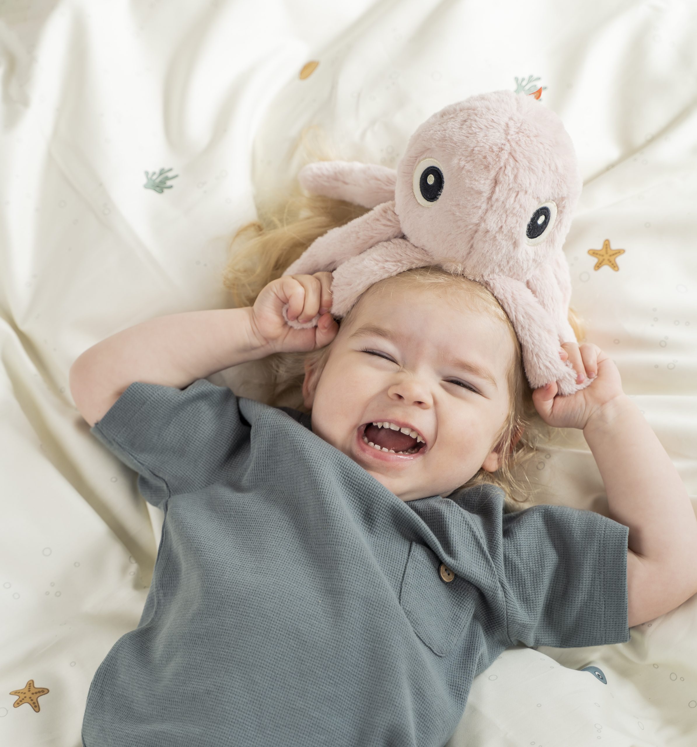 Enfant sur un lit riant avec la peluche Jelly rose poudré Done by Deer posée sur sa tête – moment ludique et joyeux