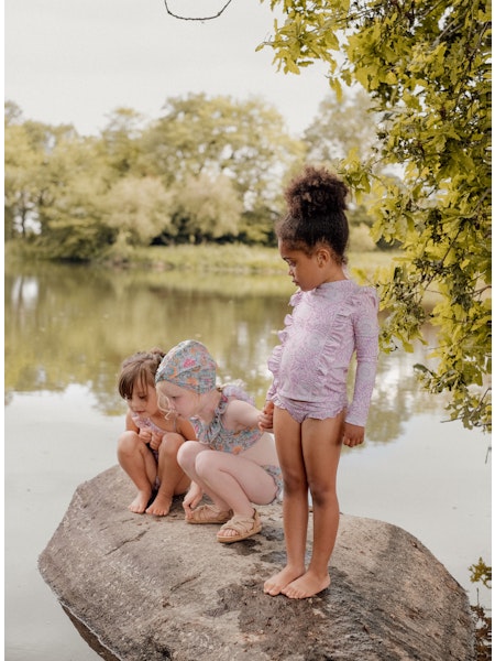 Trois enfants en maillots de bain Louise Misha, dont une fille portant l’ensemble anti-UV Yanika rose fleuri, debout sur un rocher au bord d’un étang