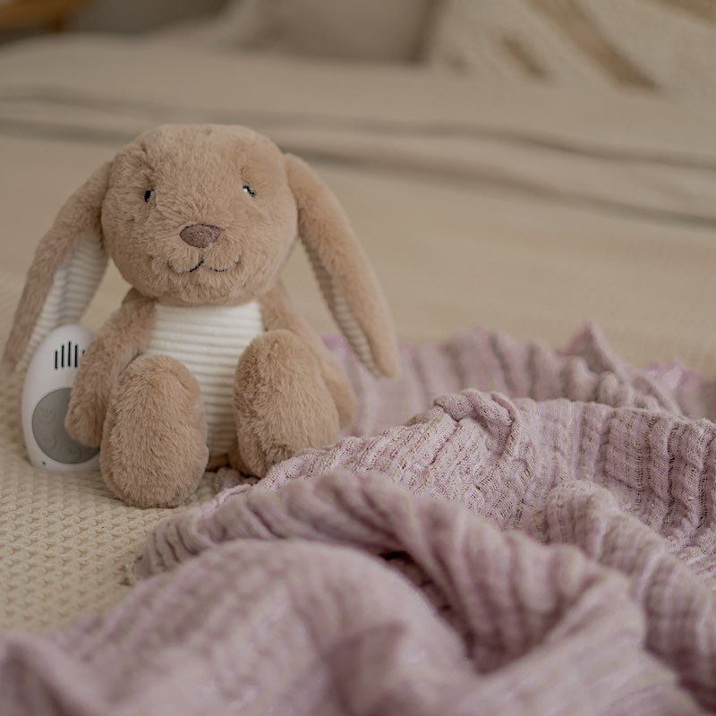 Peluche bruit blanc Milo lapin Flow Amsterdam posée sur un lit bébé avec boîtier sonore