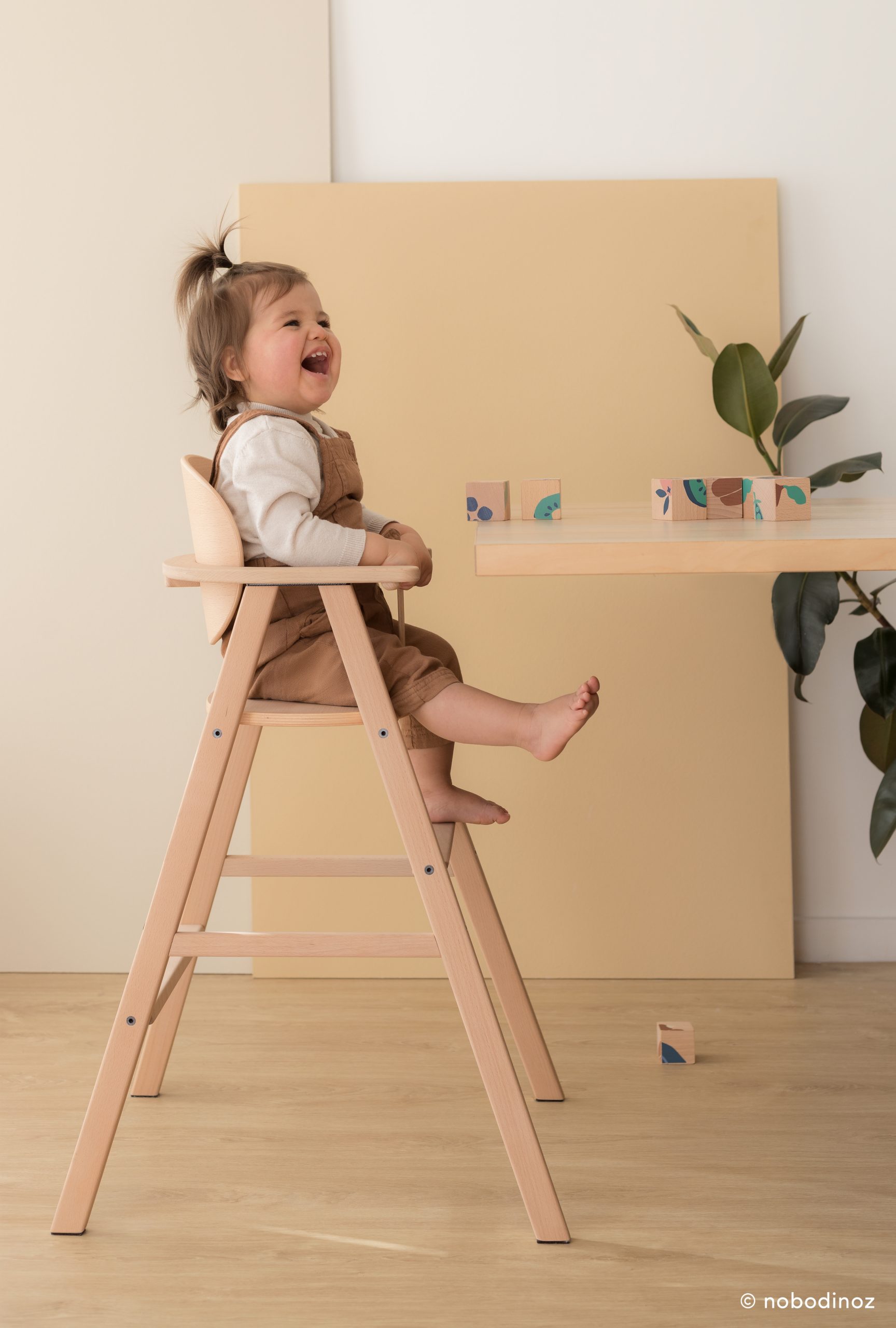 Bébé assis sur la chaise haute Nobodinoz en bois clair, jouant à table.