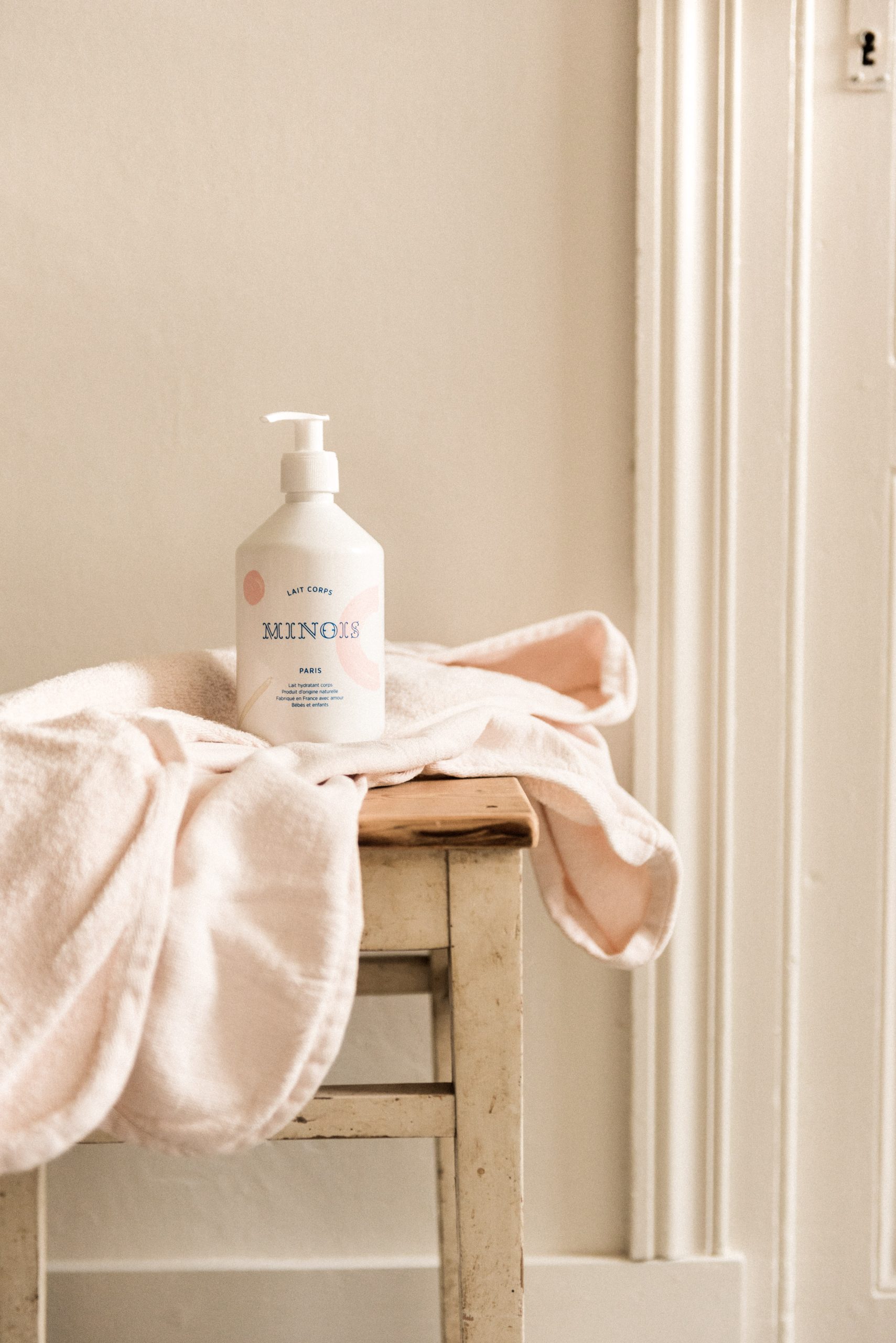 Lait hydratant bébé Minois Paris 500 ml sur chaise en bois avec serviette rose.