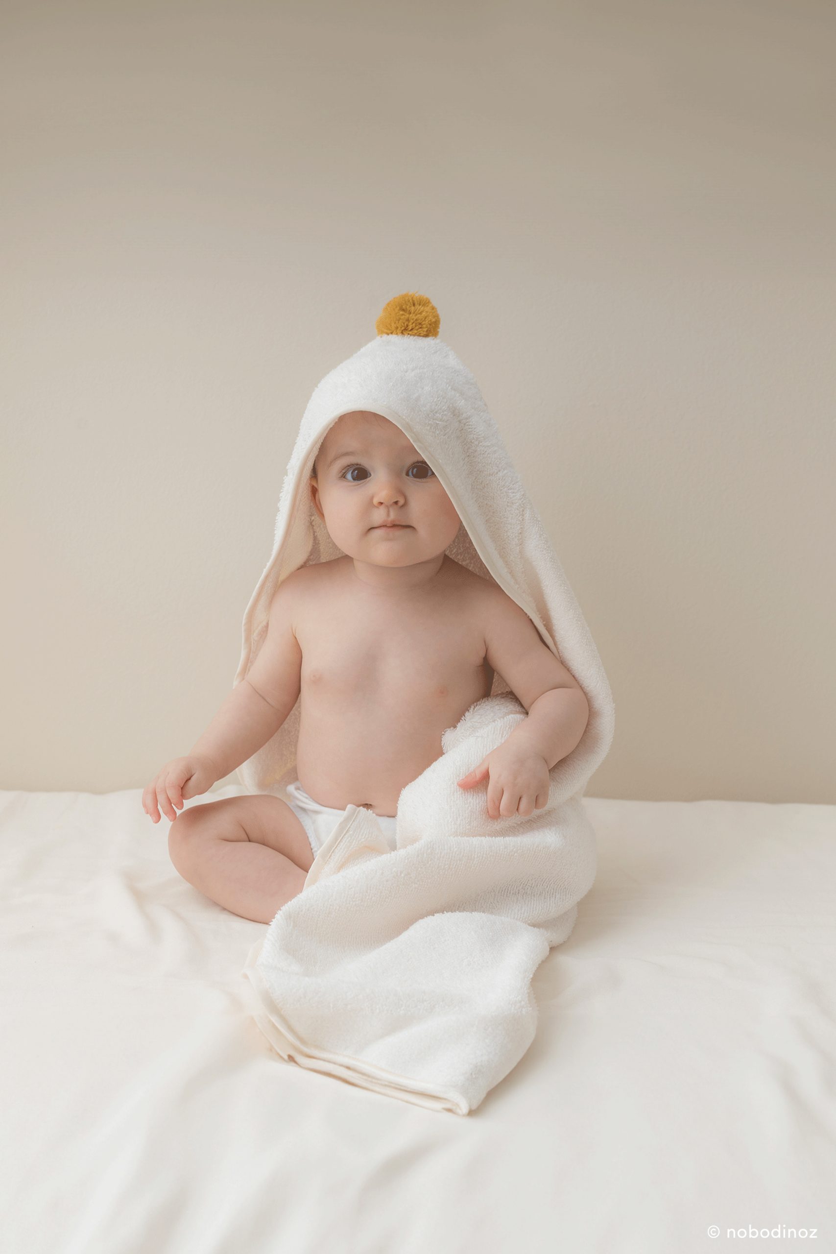 Bébé portant la cape de bain Nobodinoz en coton bio naturel avec pompon moutarde à la verticale.