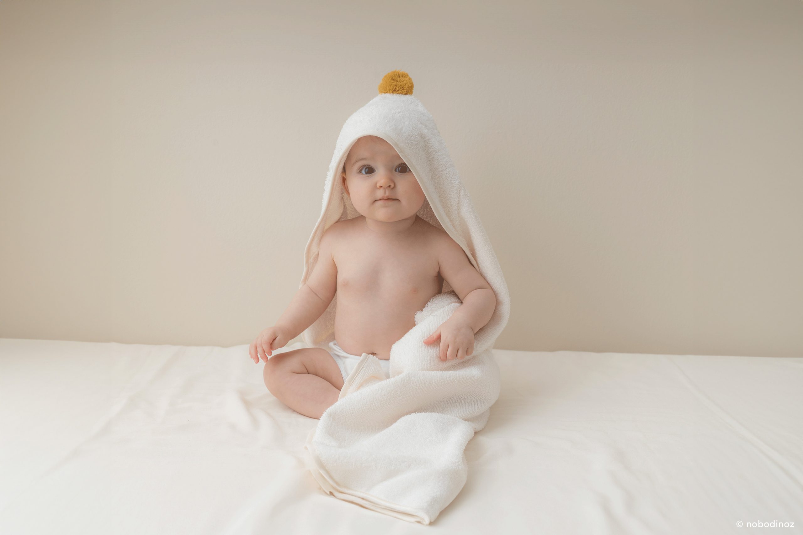 Bébé portant la cape de bain Nobodinoz en coton bio naturel avec pompon moutarde à l’horizontal.