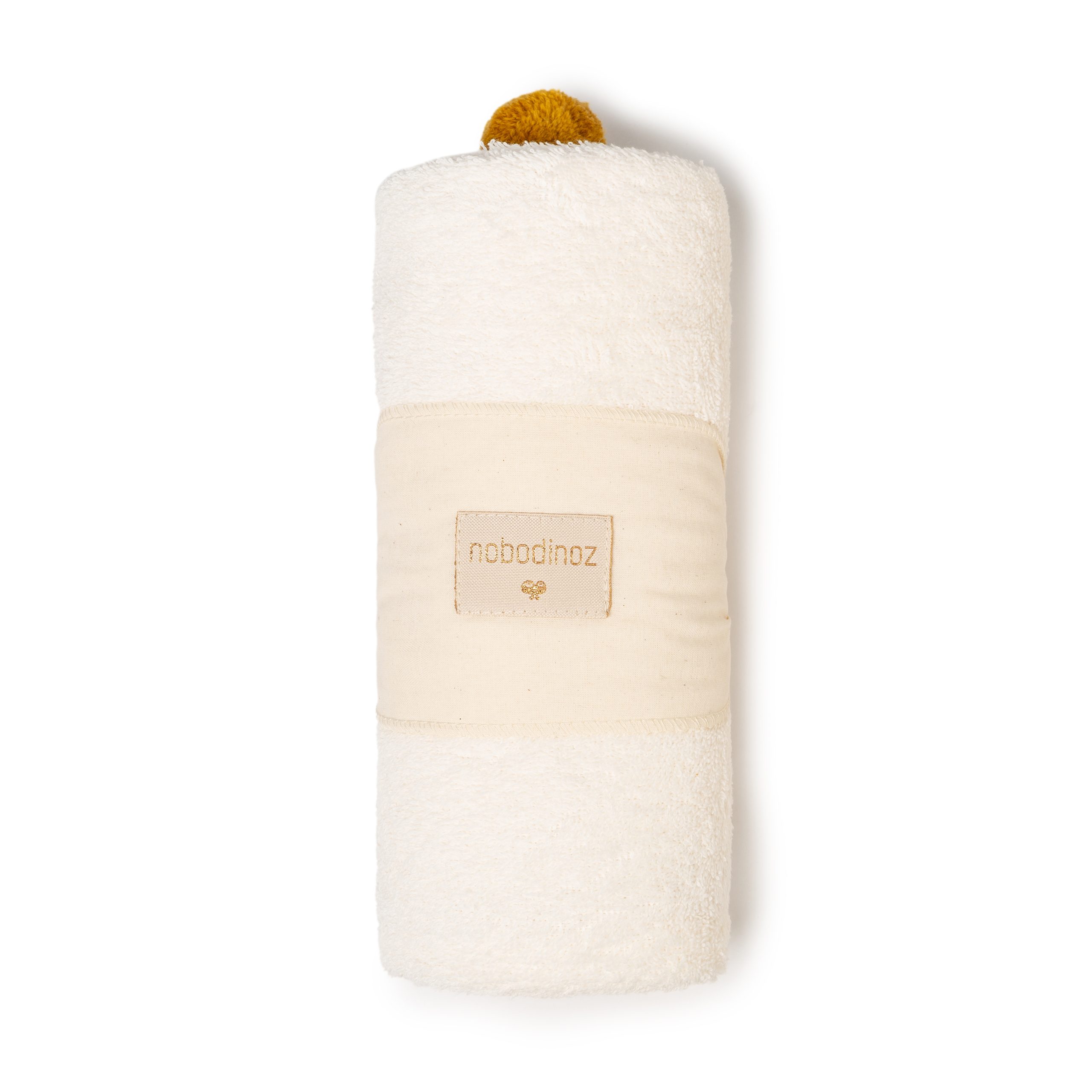 Cape de bain Nobodinoz en coton bio couleur naturel, roulée et ornée d’un pompon ocre sur le dessus.