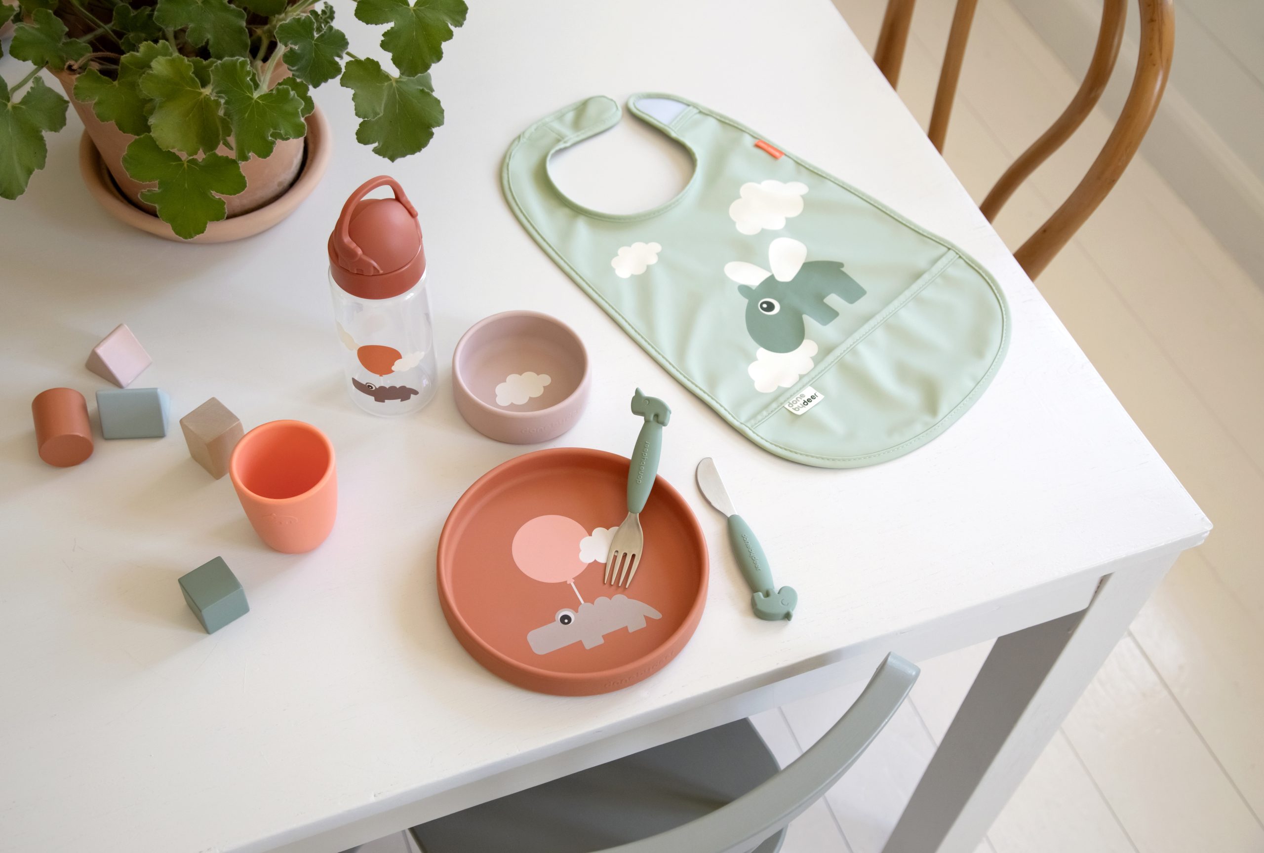 Bavoir imperméable vert Done by Deer Happy Clouds sur une table avec assiette, couverts et gobelet assortis pour bébé.