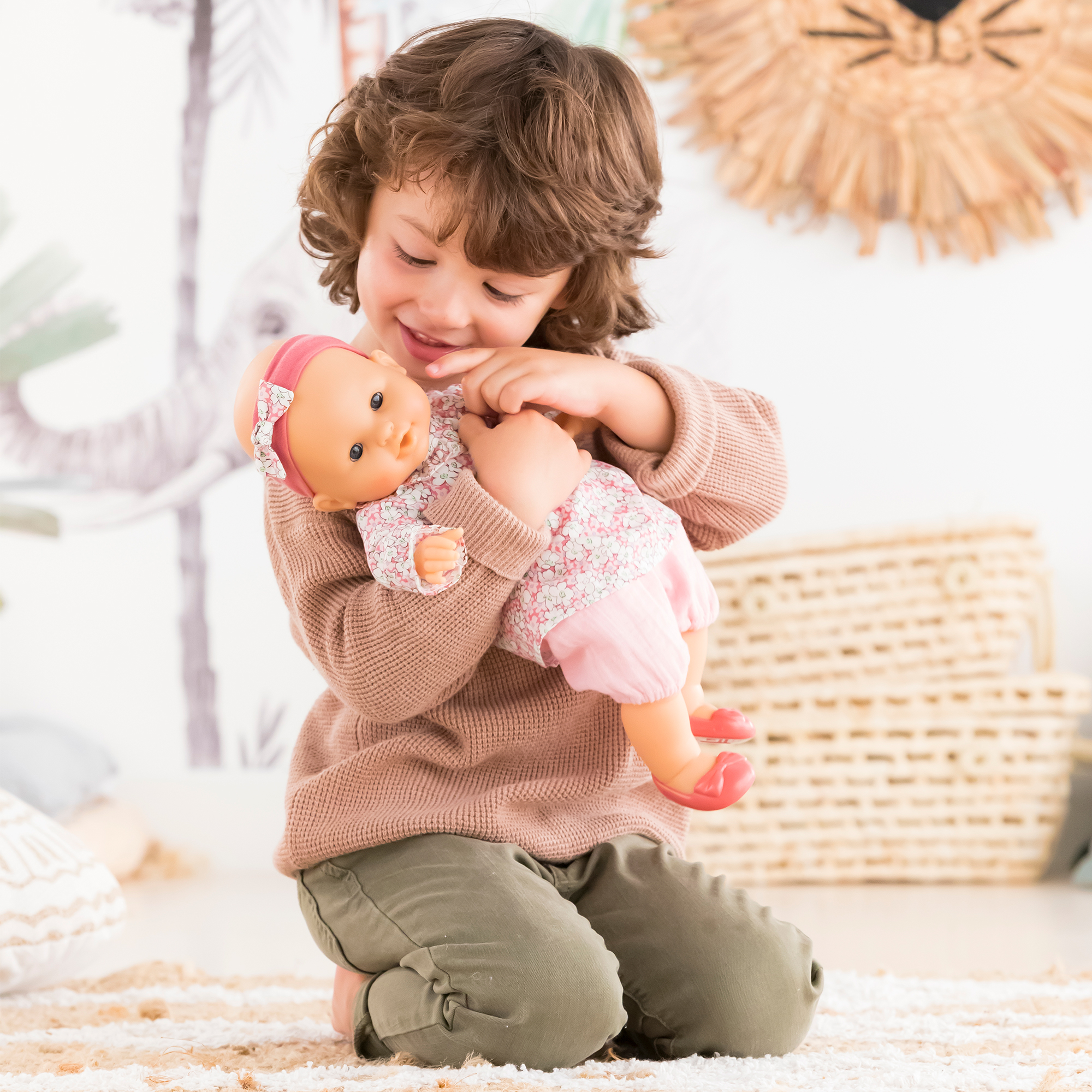 Petite fille accroupie tenant le poupon Louise 36 cm de Corolle contre elle – elle approche doucement son doigt de la joue de la poupée – moment de tendresse et d’affection
