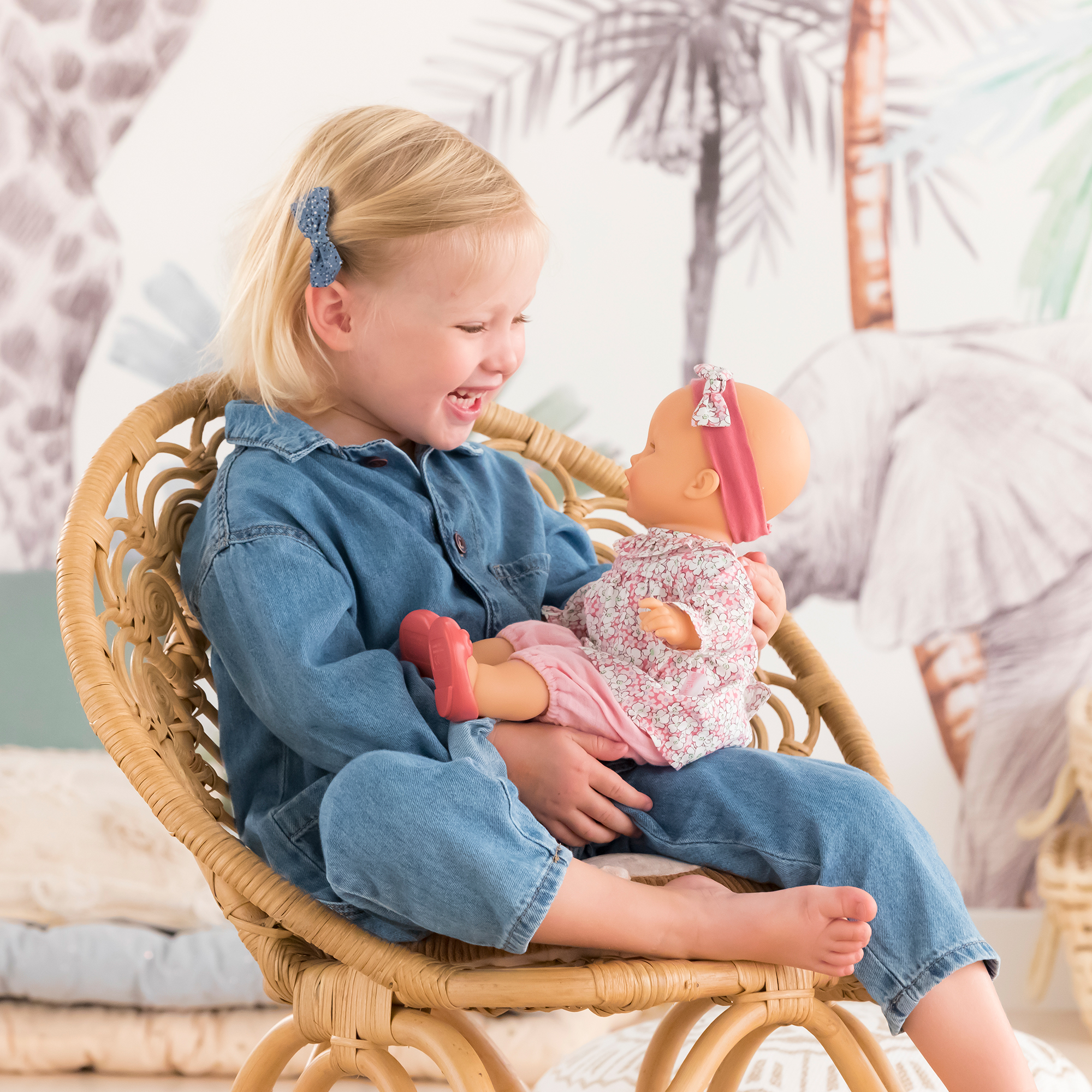 Petite fille assise sur une chaise tenant le poupon Louise 36 cm de Corolle sur ses genoux – elles se regardent et la fillette rit – moment complice de jeu d’imitation