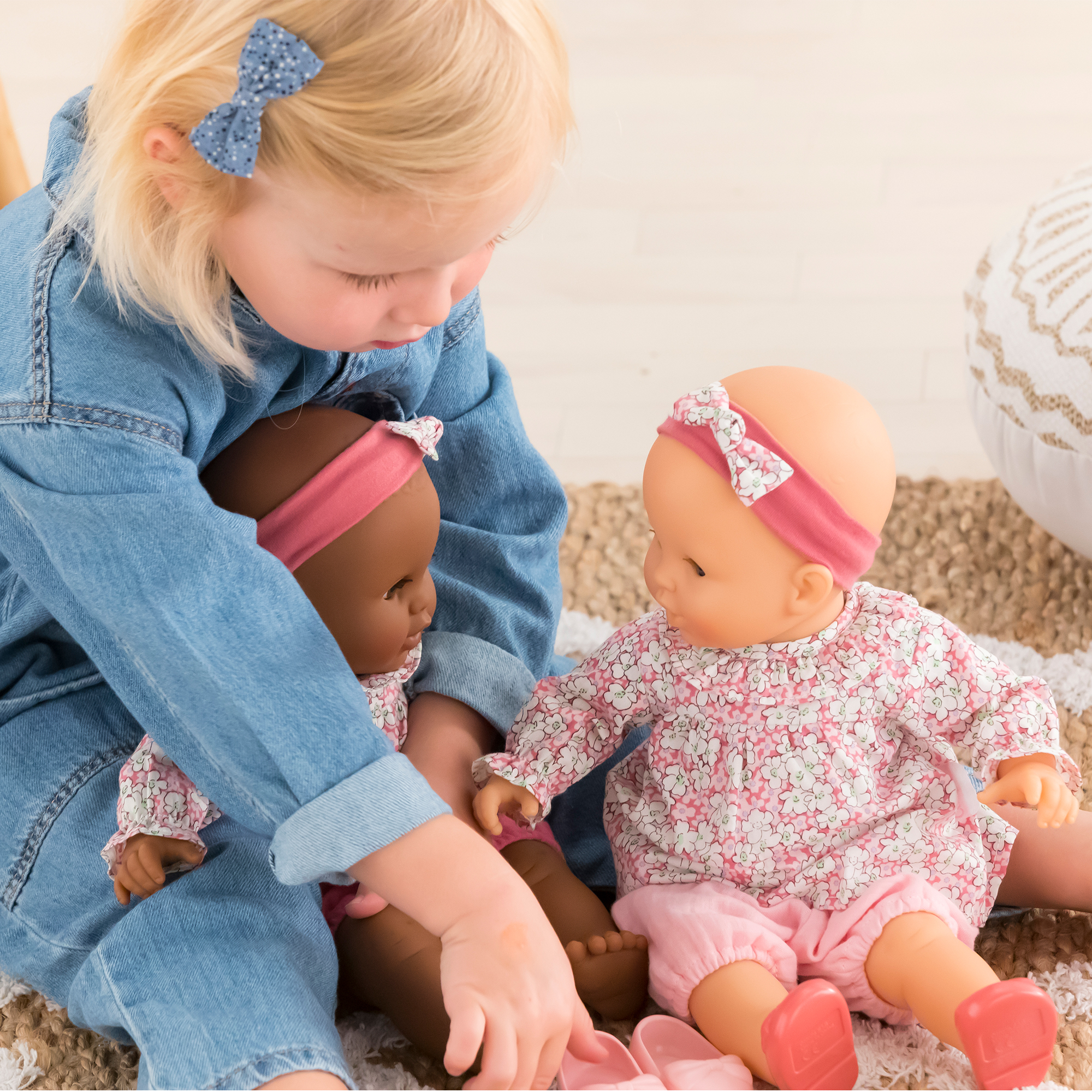 Petite fille assise jouant avec les poupons Lilou et Louise 36 cm de Corolle – Louise à sa droite et Lilou entre ses jambes pendant qu’elle remet les chaussures – scène de soin et de jeu d’imitation