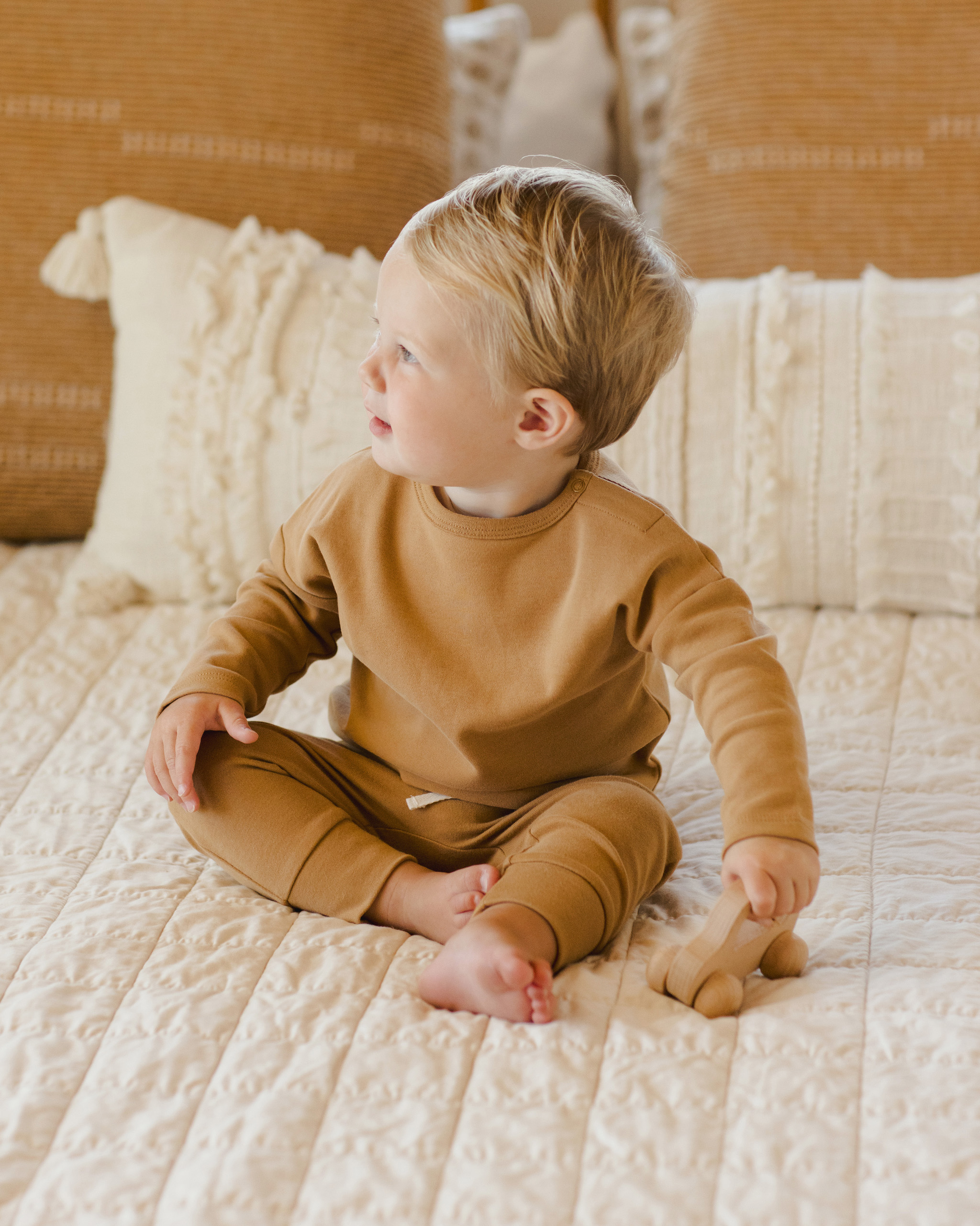 Enfant de 2 ans assis sur un lit, portant un ensemble Quincy Maé en coton bio ocre : t-shirt manches longues et pantalon à taille élastique