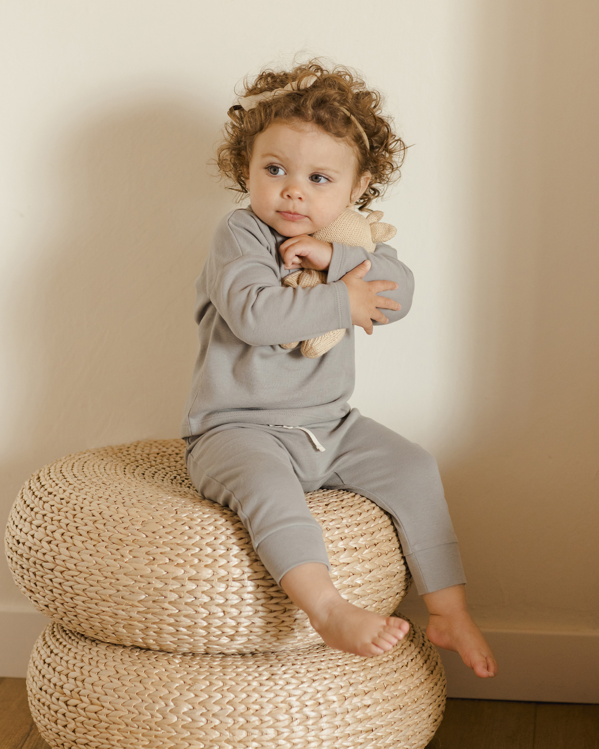Bébé fille assise sur un pouf, portant un pantalon gris bleu Quincy Mae et serrant une peluche dans ses bras