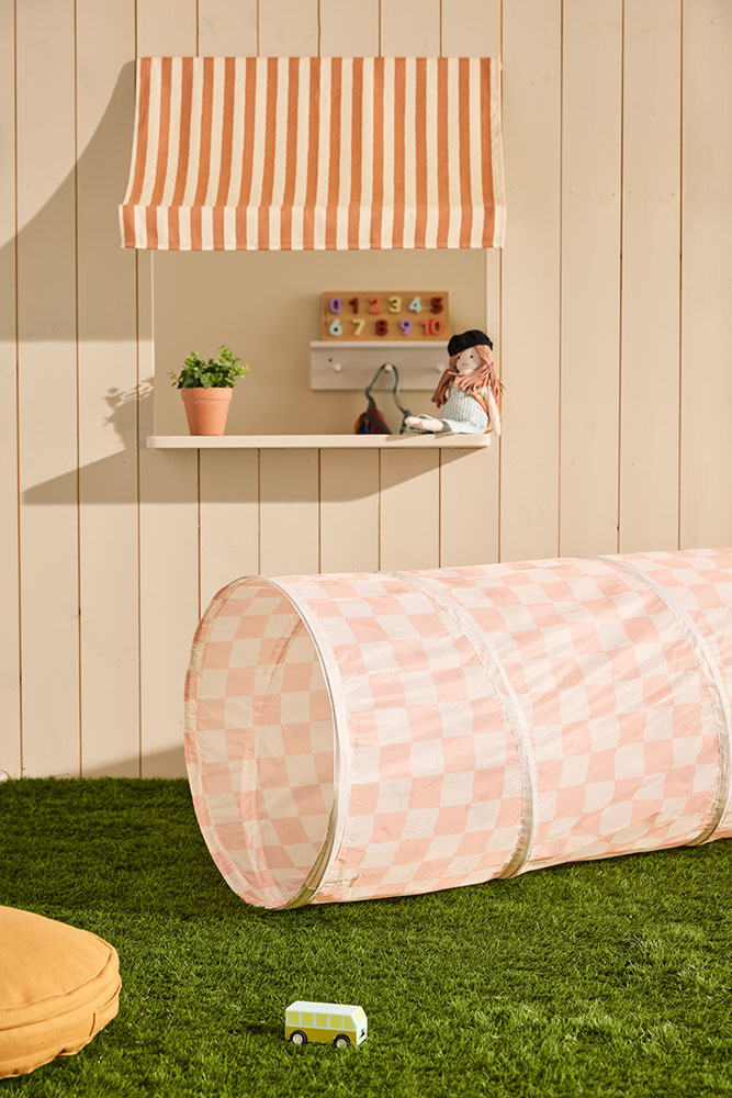 Tunnel de jeu bébé en tissu à carreaux abricot, posé sur de l’herbe, devant une cabane en bois – Kid’s Concept