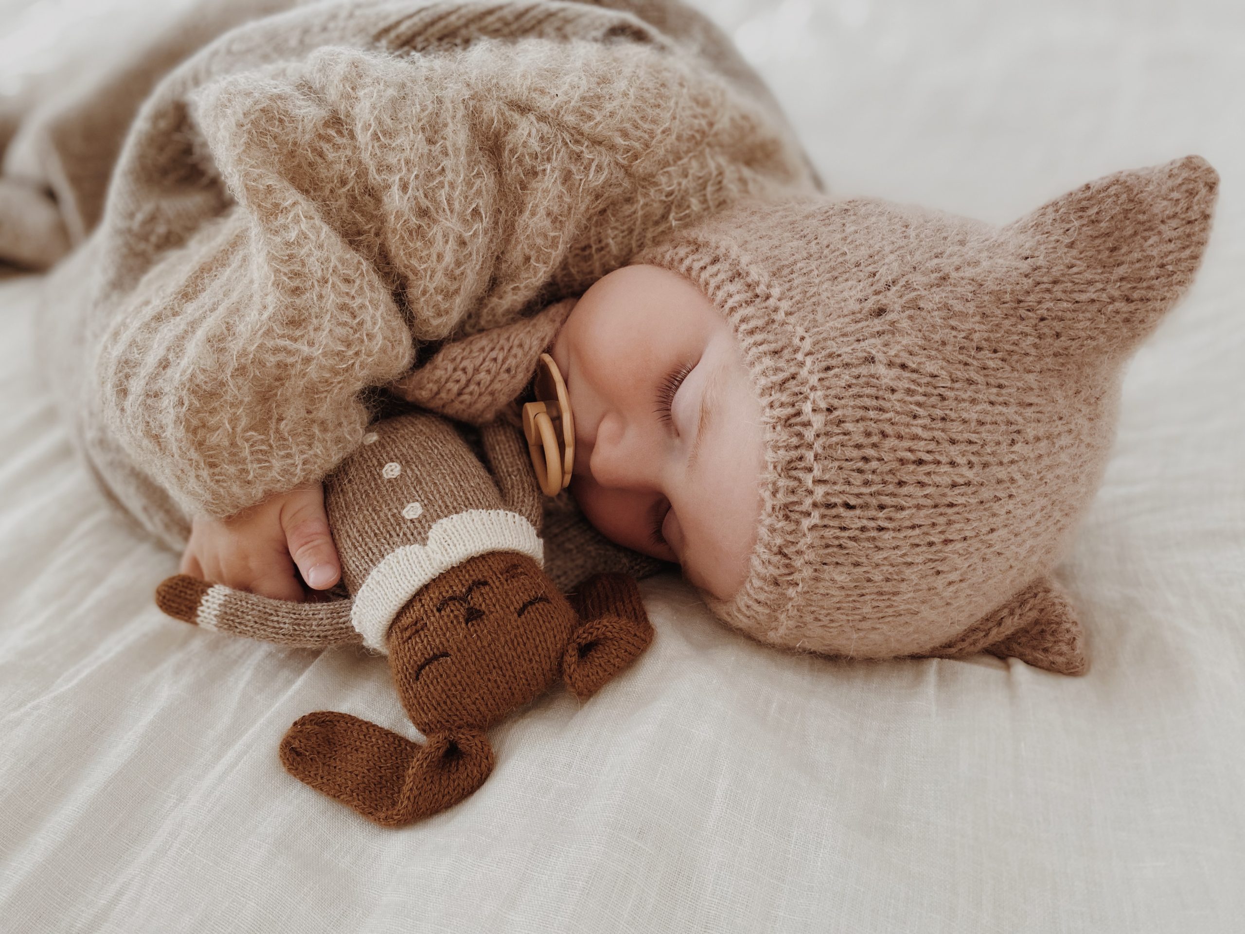 Bébé dormant sur le ventre tenant le doudou Lapin Combinaison avoine Main Sauvage dans son bras – sur un lit