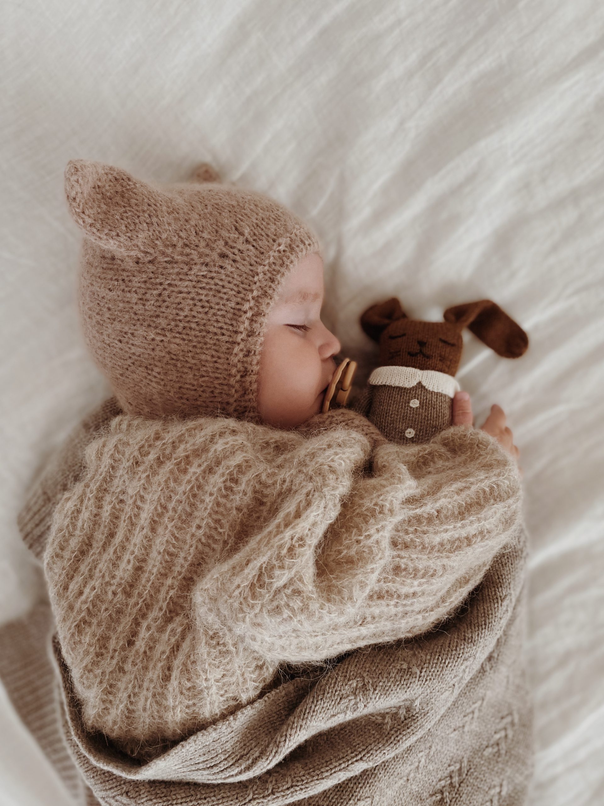 Vue du dessus d’un bébé dormant sur le ventre tenant le doudou Lapin Combinaison avoine Main Sauvage – sur un lit