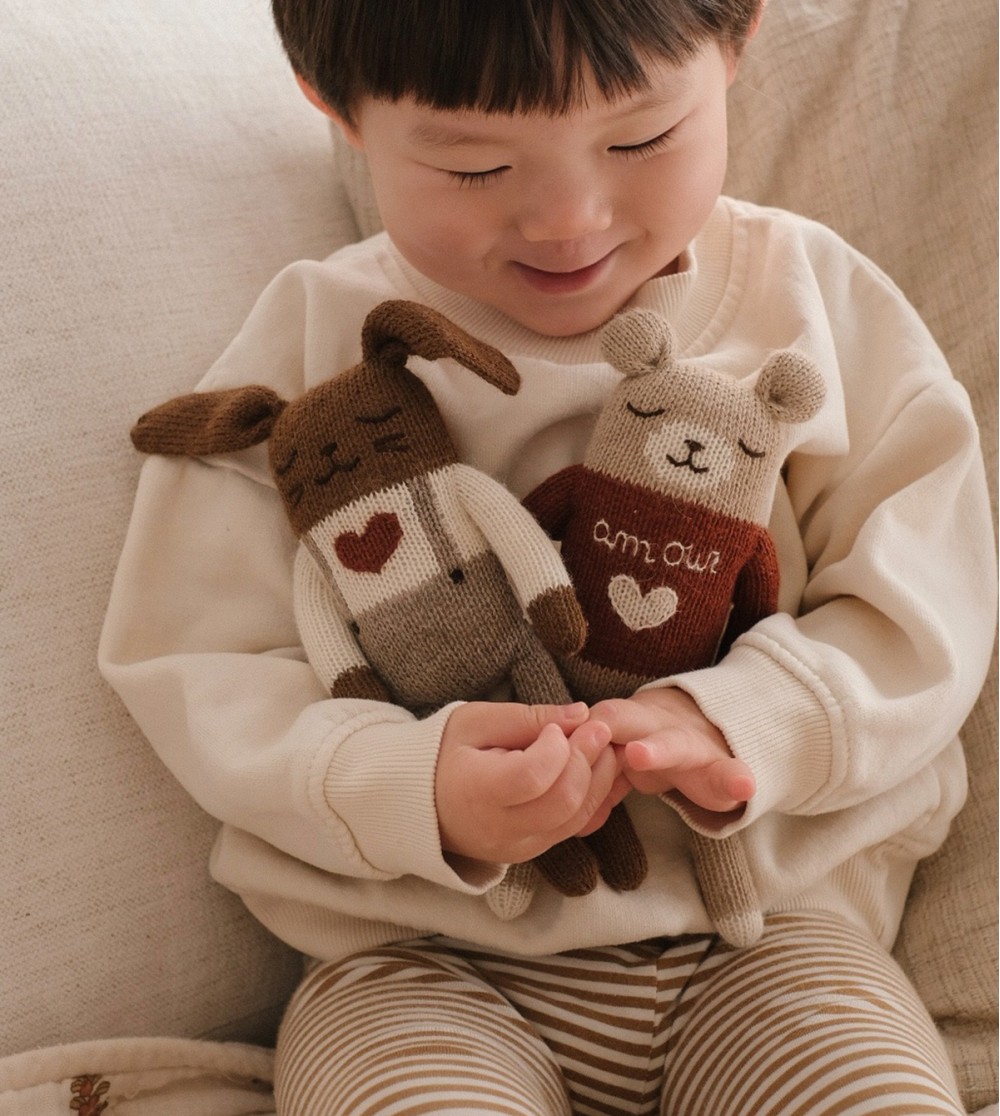 Enfant assis sur un canapé tenant le doudou Lapin Amour Main Sauvage, avec le doudou Ourson Amour à ses côtés – moment de joie