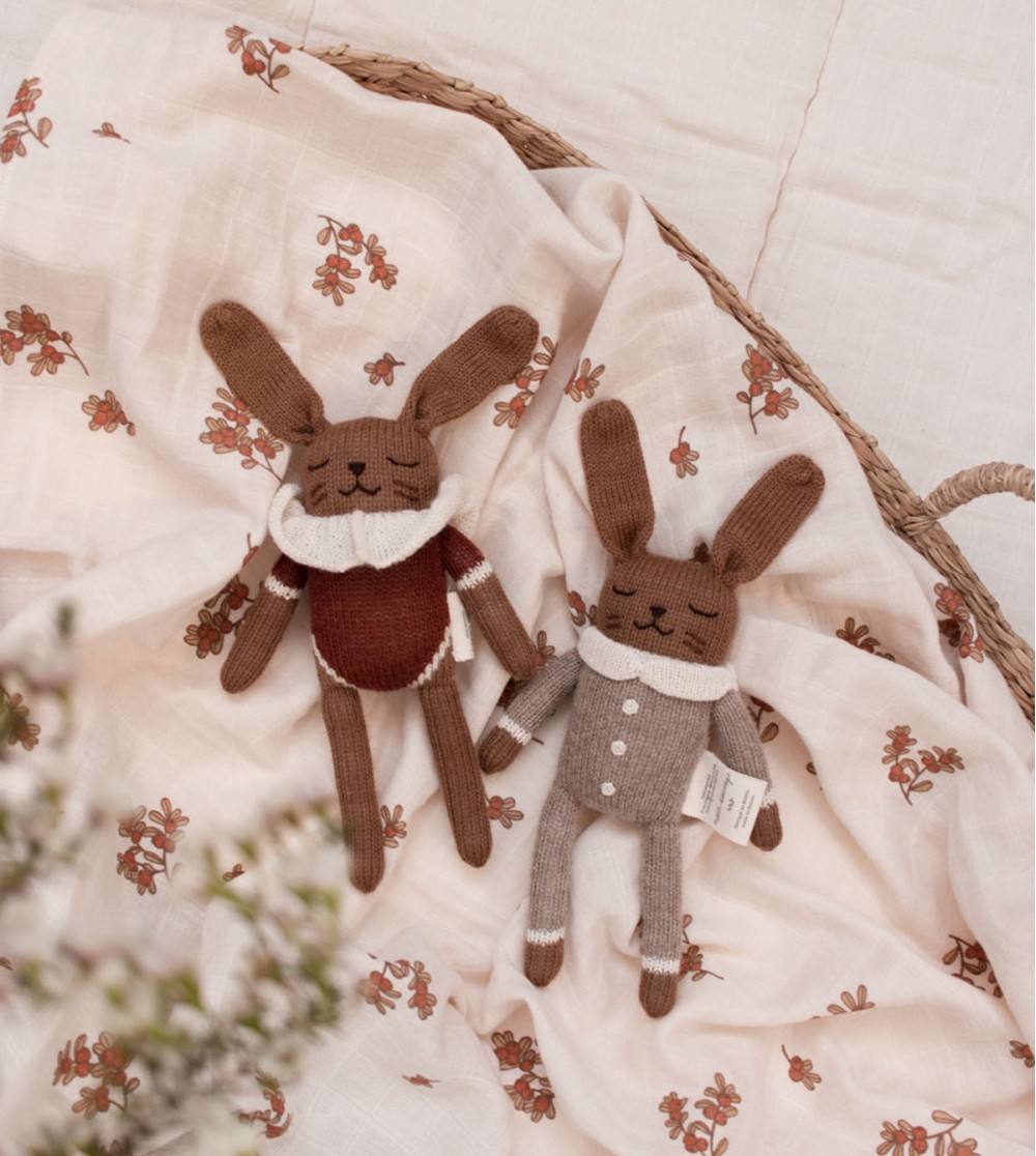Doudou Lapin Combinaison avoine Main Sauvage et un autre doudou lapin posés dans un couffin sur un drap fleuri – vue du dessus