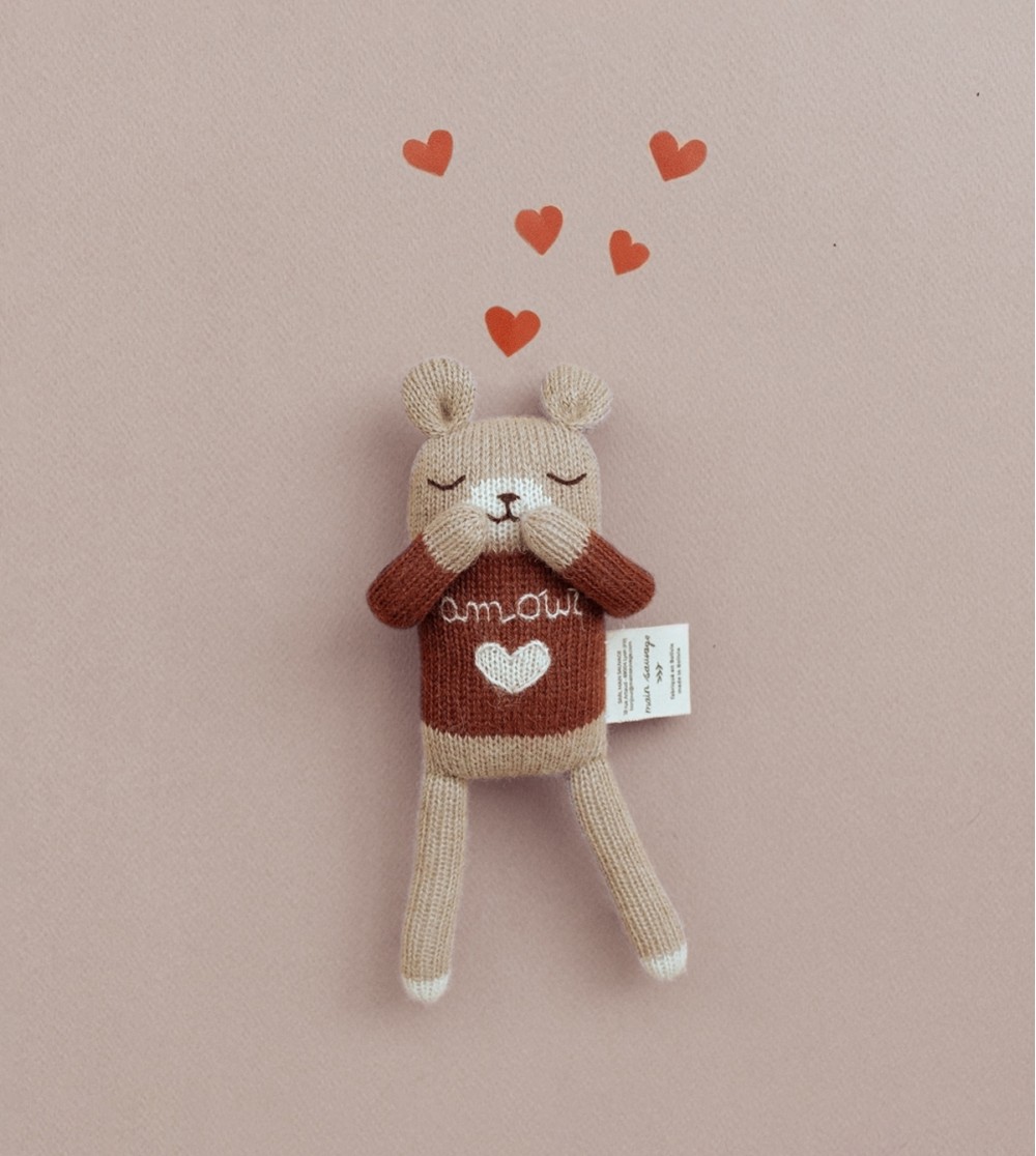 Doudou Ourson Amour Main Sauvage posé sur fond rosé, bras levés vers sa bouche dans une posture coquine, avec des petits cœurs rouges au-dessus de sa tête