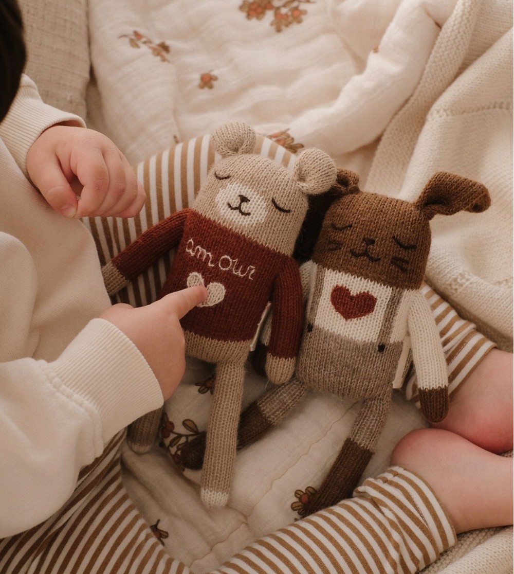 Enfant assis en tailleur sur un lit pointant du doigt le doudou Ourson Amour Main Sauvage posé sur ses jambes à côté du doudou Lapin Amour