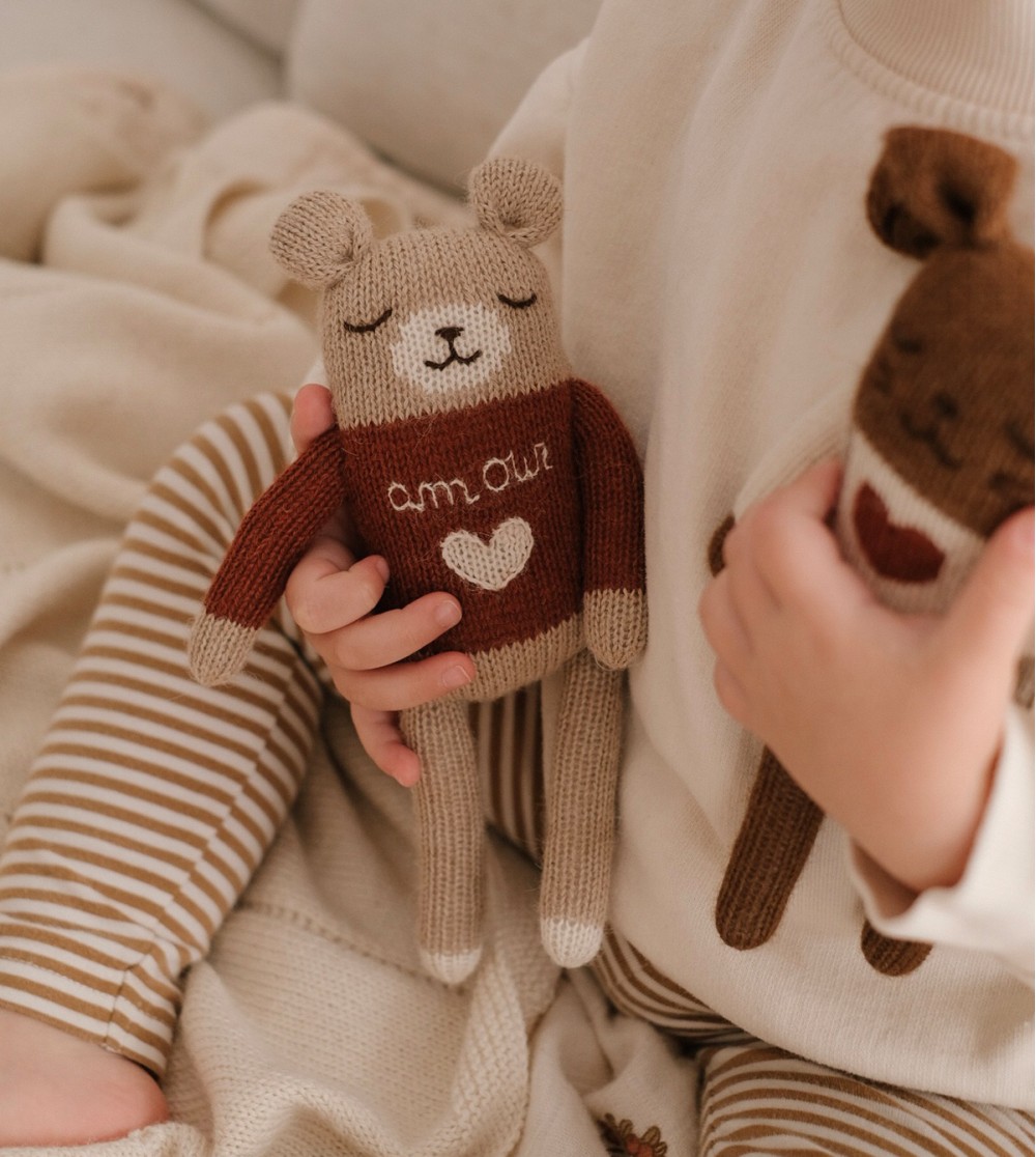 Enfant assis tenant le doudou Ourson Amour Main Sauvage contre sa poitrine et le doudou Lapin Amour dans l’autre main – vue de profil