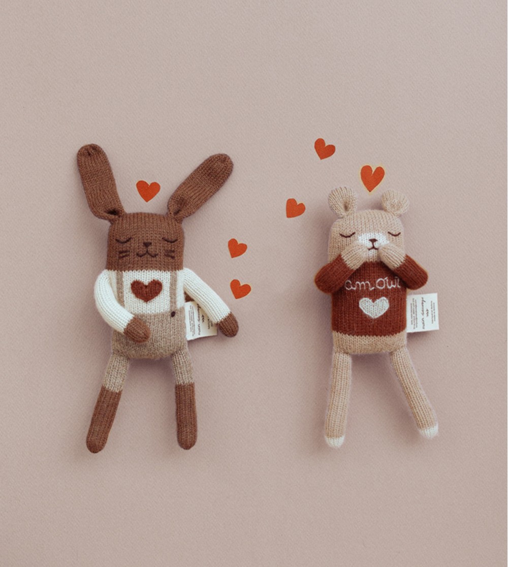 Doudous Ourson Amour et Lapin Amour Main Sauvage posés sur un fond rose, avec des cœurs entre eux – l’ourson dans une posture coquine, le lapin bras croisé sur le buste – ambiance tendre