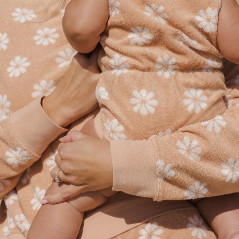 Bébé en combinaison éponge sans manches Rylee + Cru à fleurs blanches sur fond rose pamplemousse, porté dans les bras d’un adulte.
