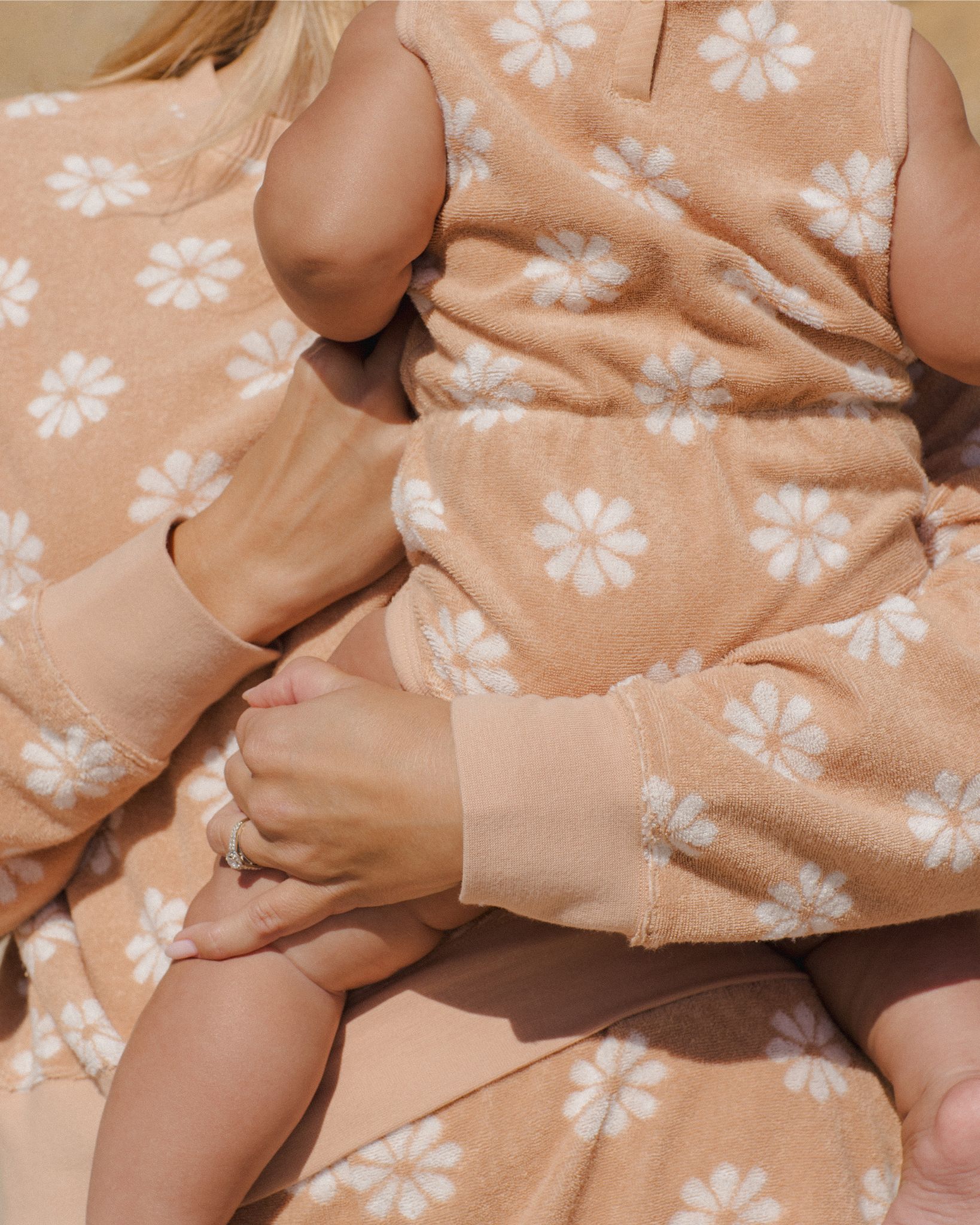 Bébé en combinaison éponge sans manches Rylee + Cru à fleurs blanches sur fond rose pamplemousse, porté dans les bras d’un adulte.