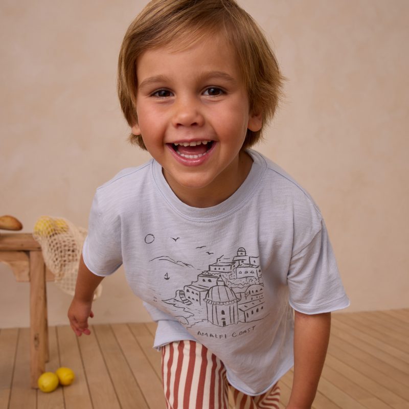 Enfant souriant portant un t-shirt Rylee + Cru bleu clair avec imprimé Amalfi Coast