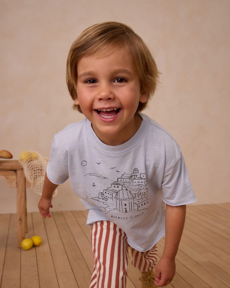 Enfant souriant portant un t-shirt Rylee + Cru bleu clair avec imprimé Amalfi Coast