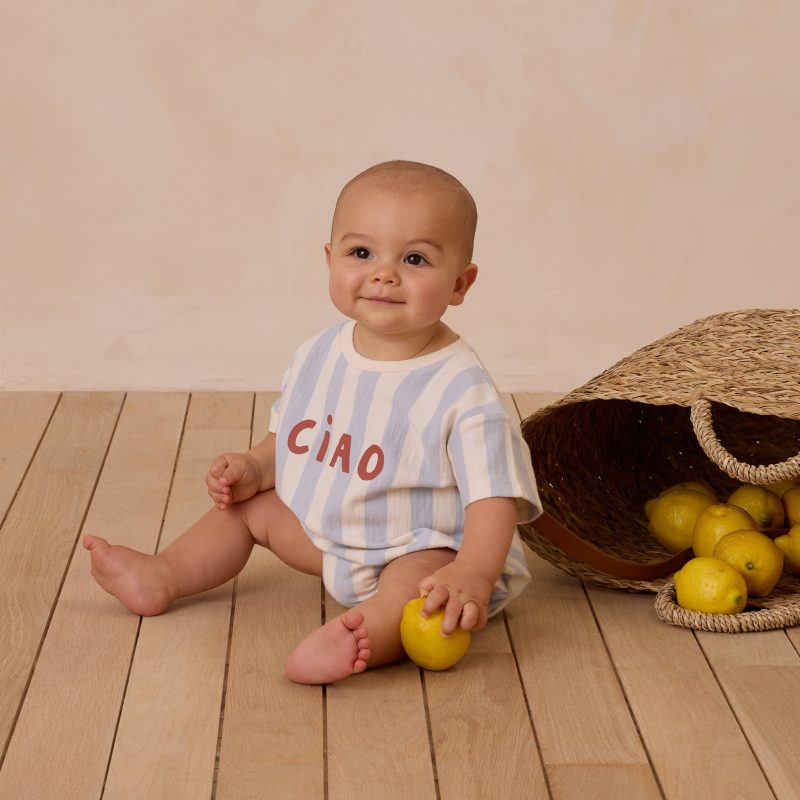 Bébé assis portant une combinaison à rayures bleu clair Rylee + Cru avec l’inscription "Ciao", assis près d’un panier de citrons.