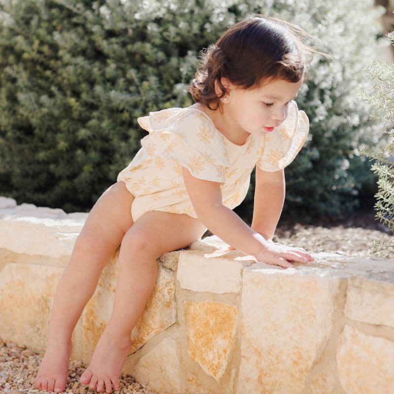 Petite fille portant une combinaison Roxy Rylee + Cru à imprimé marguerites, avec manches volantées et tissu en coton naturel.