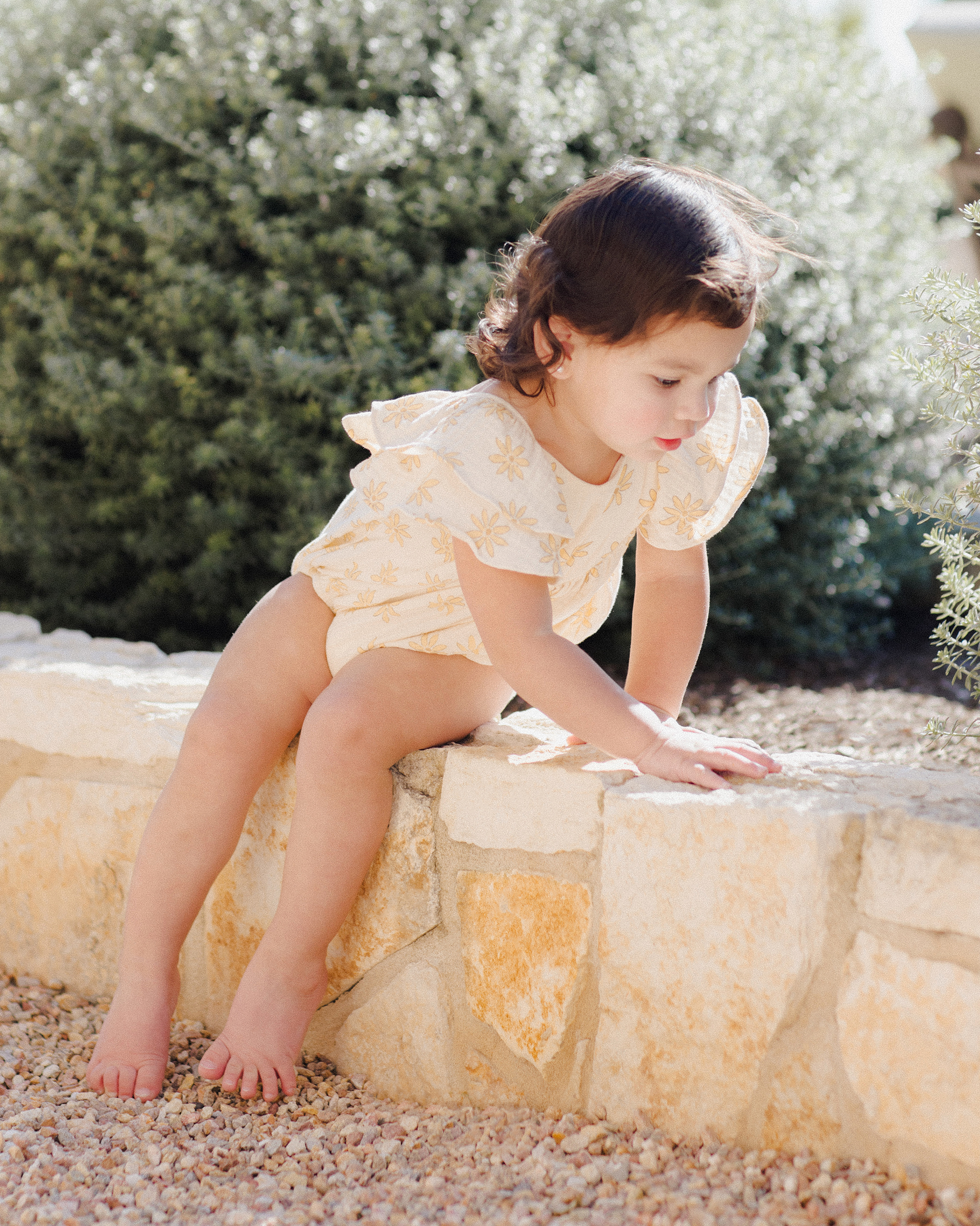 Petite fille portant une combinaison Roxy Rylee + Cru à imprimé marguerites, avec manches volantées et tissu en coton naturel.