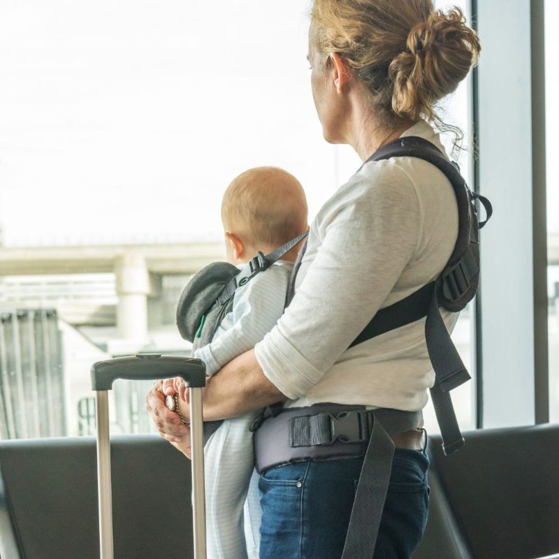 Maman portant son bébé en porte-bébé à l’aéroport, debout près d’une valise, en train de regarder par la fenêtre