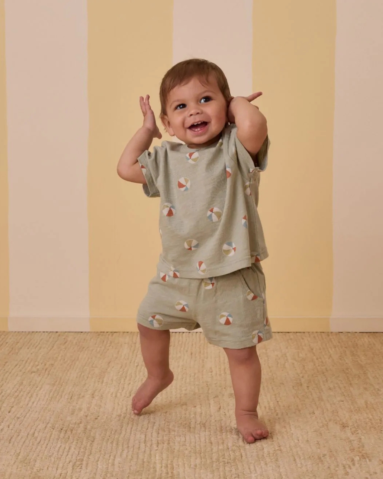 Bébé souriant portant un ensemble vert sauge à imprimé ballons de plage Rylee + Cru, composé d’un T-shirt et d’un short.