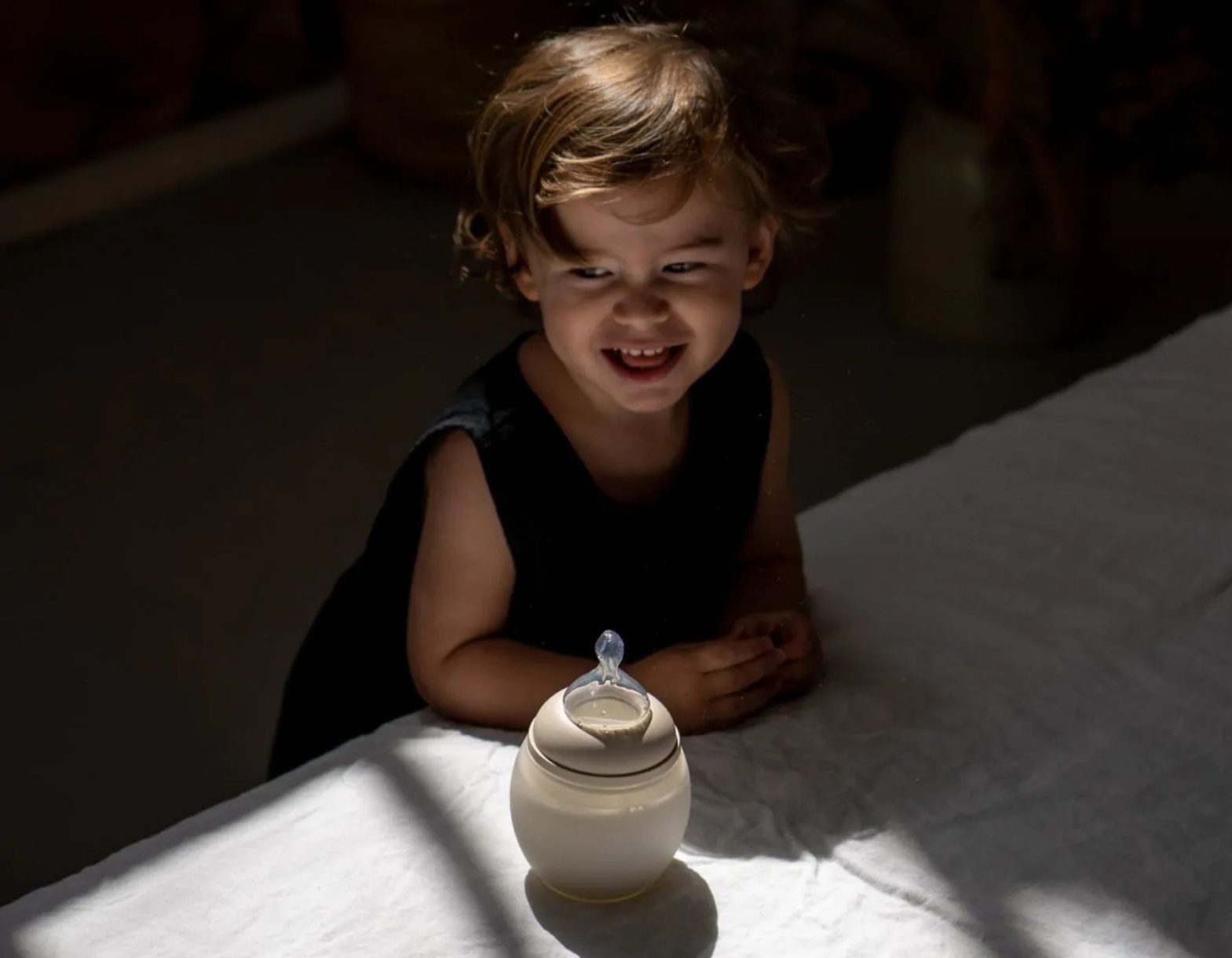 Enfant souriant avec un biberon Elhée sur une table baignée de lumière